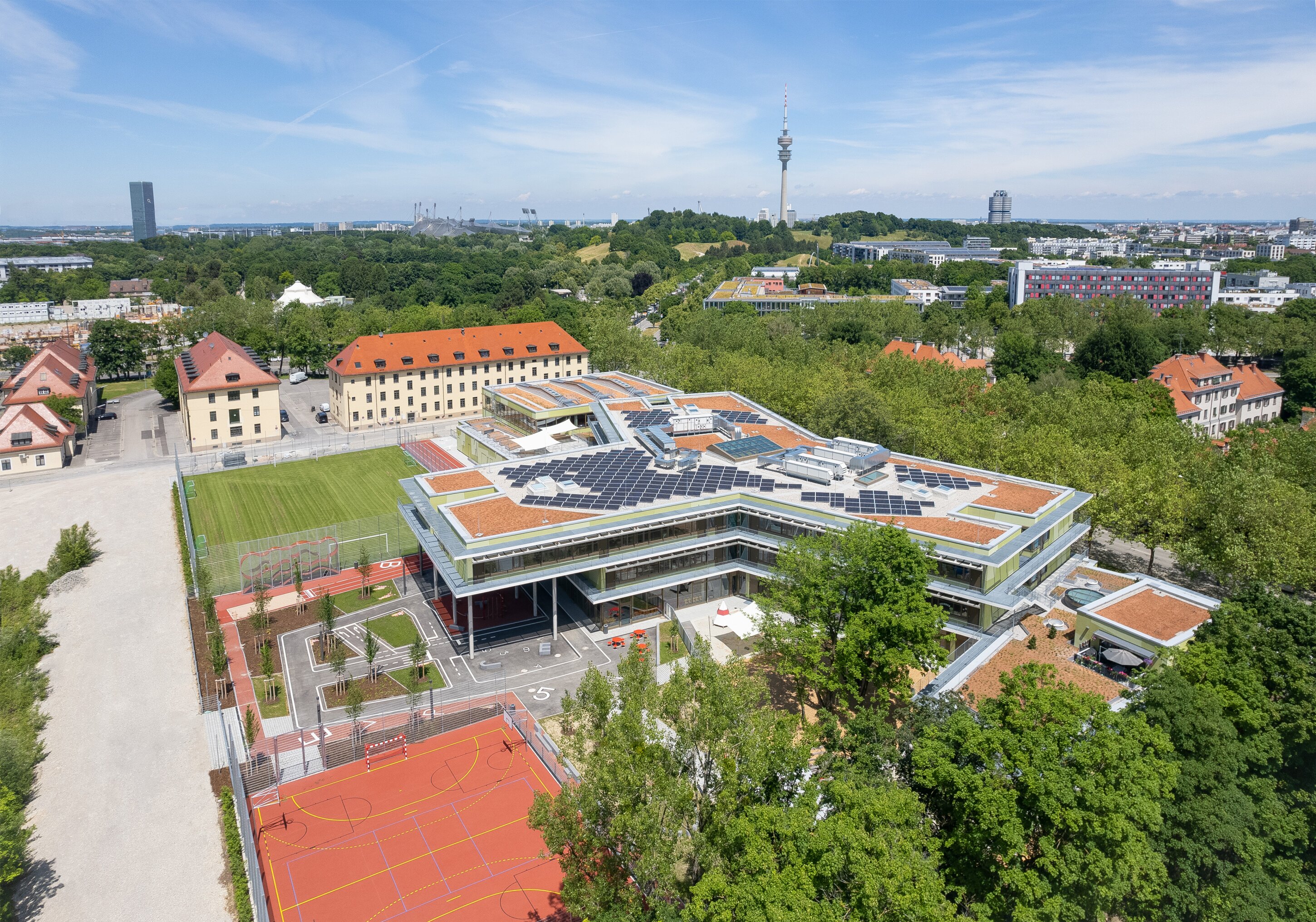Project by Behnisch Architekturbuero, Primaryschool Infanteriestra&szlig;e. Aerial view of a modern school complex with solar panels, sports fields and courts, surrounded by trees and city buildings.