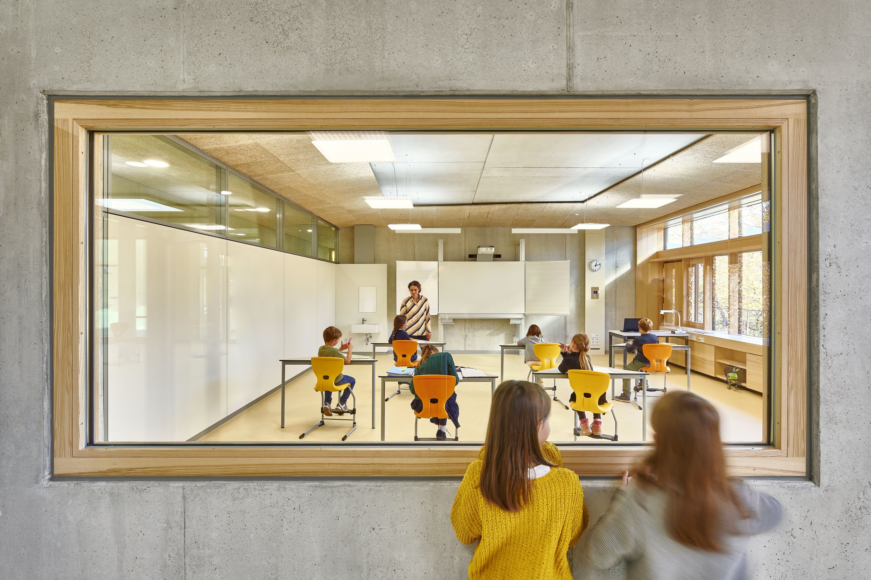 Project by Behnisch Architekturbuero, Primaryschool Infanteriestra&szlig;e. View through a large window into a classroom where a teacher instructs seated children, while two students watch from the corridor outside.