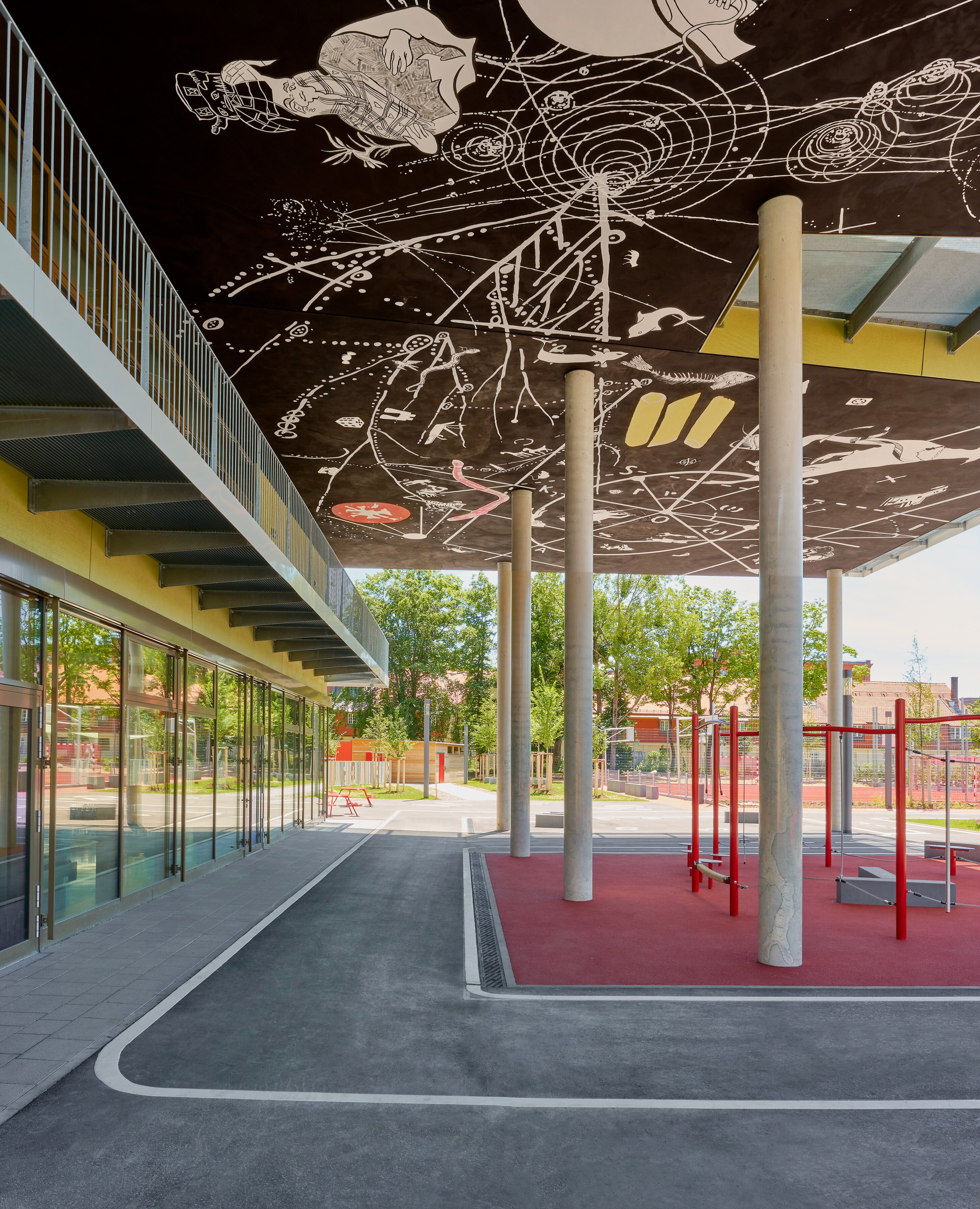 Project by Behnisch Architekturbuero, Primaryschool Infanteriestra&szlig;e. Covered outdoor area with columns and a ceiling mural, next to a building and a playground with red equipment on a rubber surface.