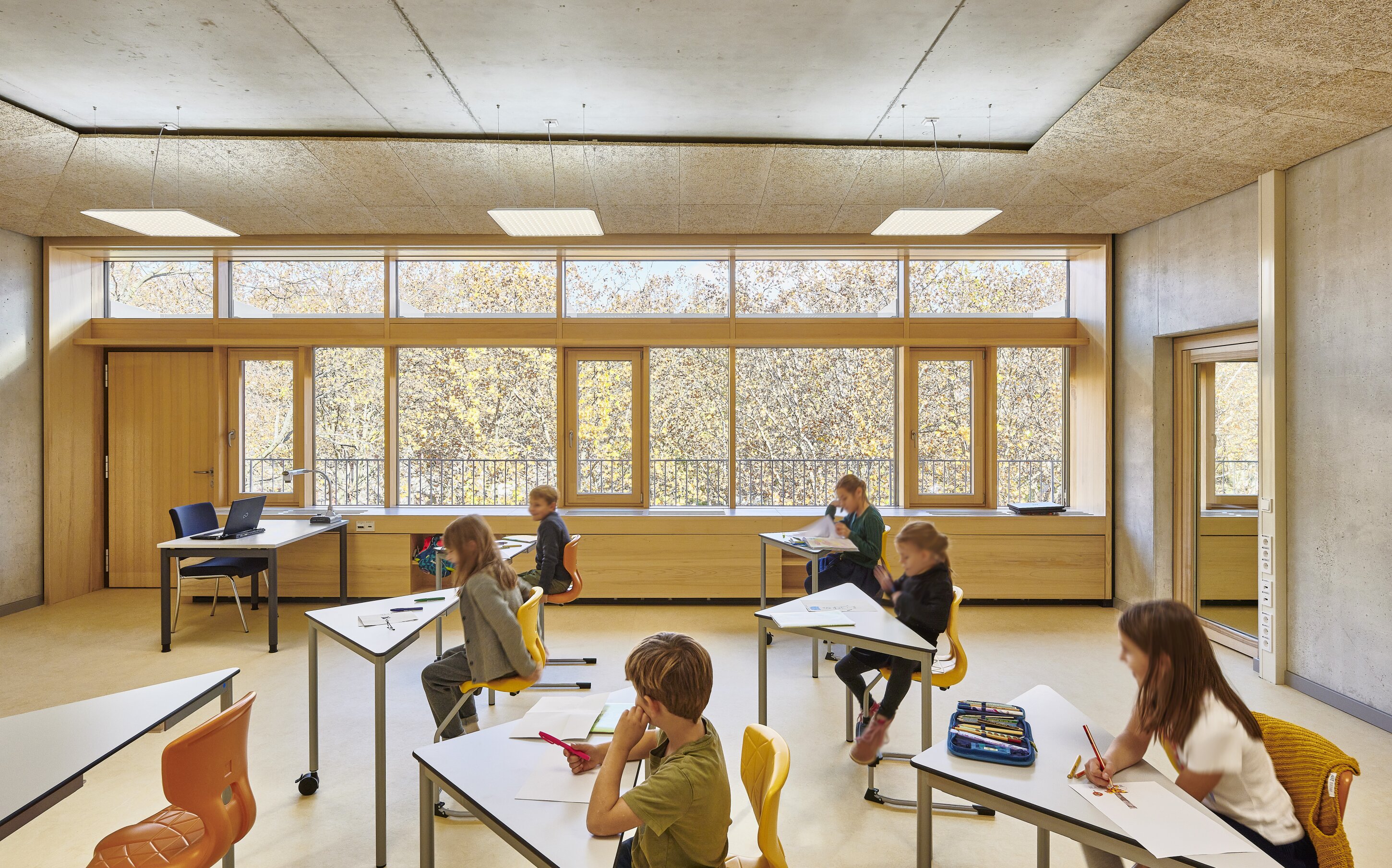 Project by Behnisch Architekturbuero, Primaryschool Infanteriestra&szlig;e. Classroom with children seated at individual desks working, large windows along the wall, and a teacher&rsquo;s desk with a laptop at the front.