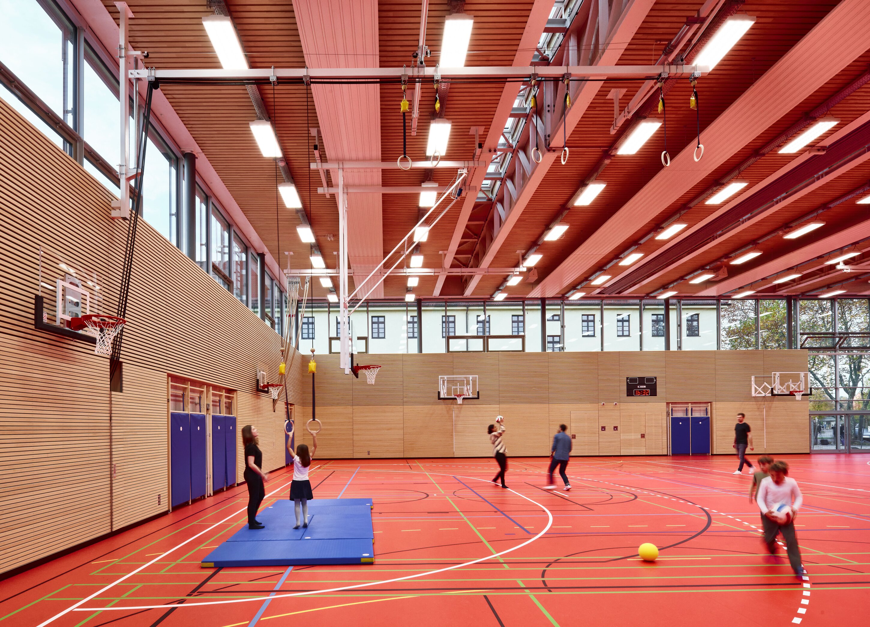 Project by Behnisch Architekturbuero, Primaryschool Infanteriestra&szlig;e. Indoor gymnasium with red floor, basketball hoops and rings, where children and adults are playing and exercising.