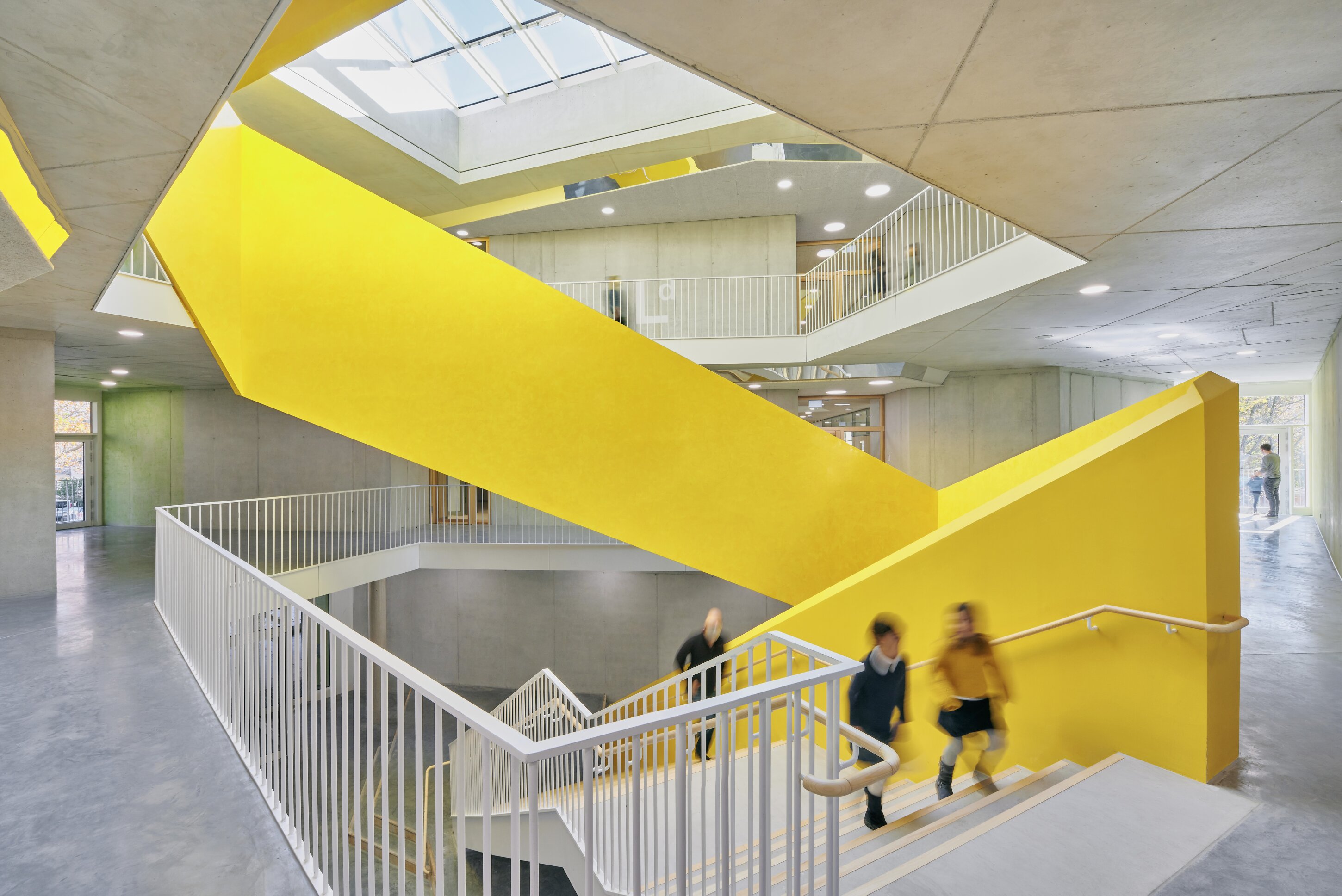 Project by Behnisch Architekturbuero, Primaryschool Infanteriestra&szlig;e. Interior atrium with a bright yellow staircase, concrete walls, skylight above, and people moving on stairs and upper walkways.