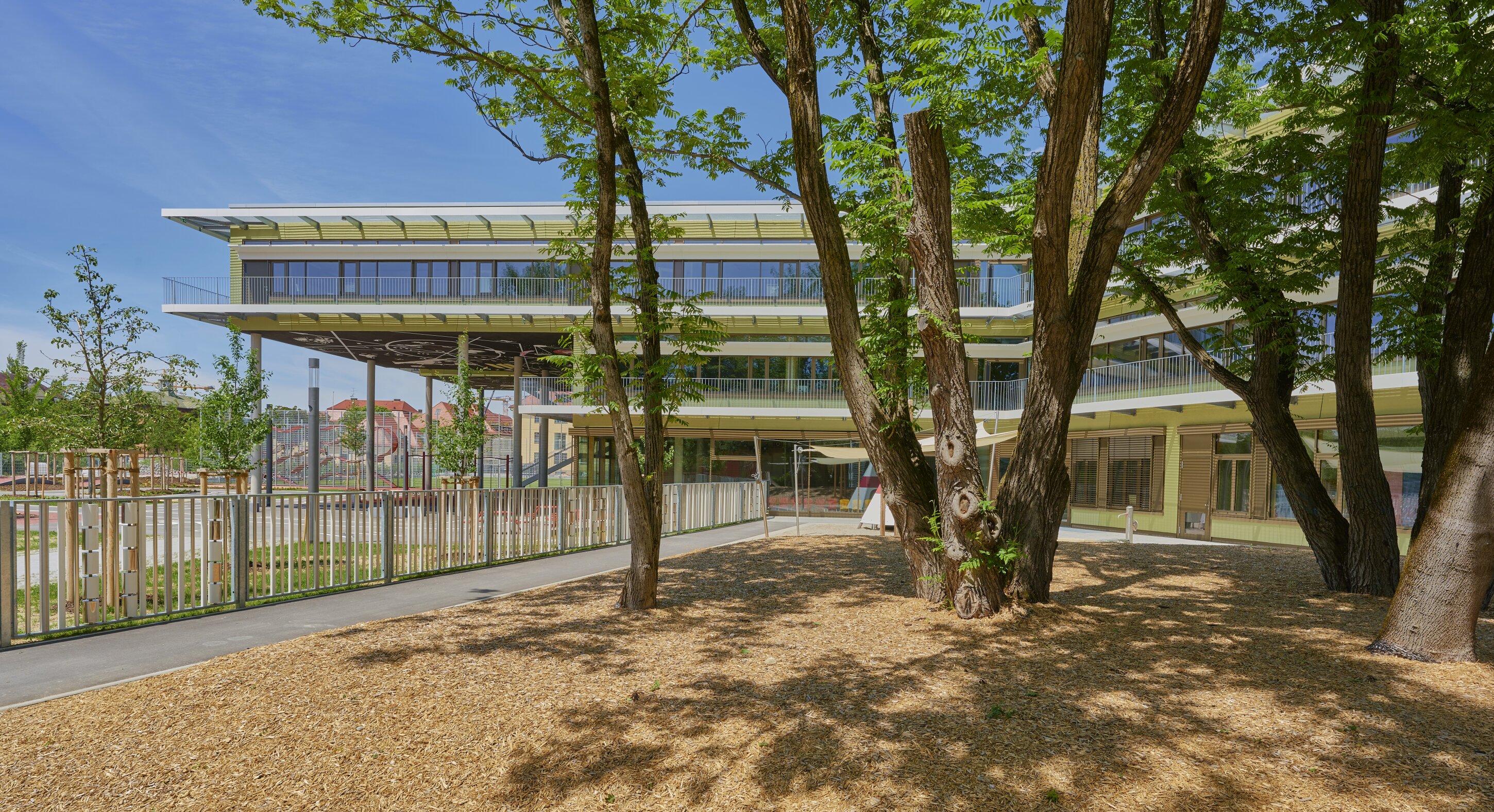 Project by Behnisch Architekturbuero, Primaryschool Infanteriestra&szlig;e. School courtyard with trees in the foreground, fenced play areas, and a modern building with balconies and shaded walkways in the background.