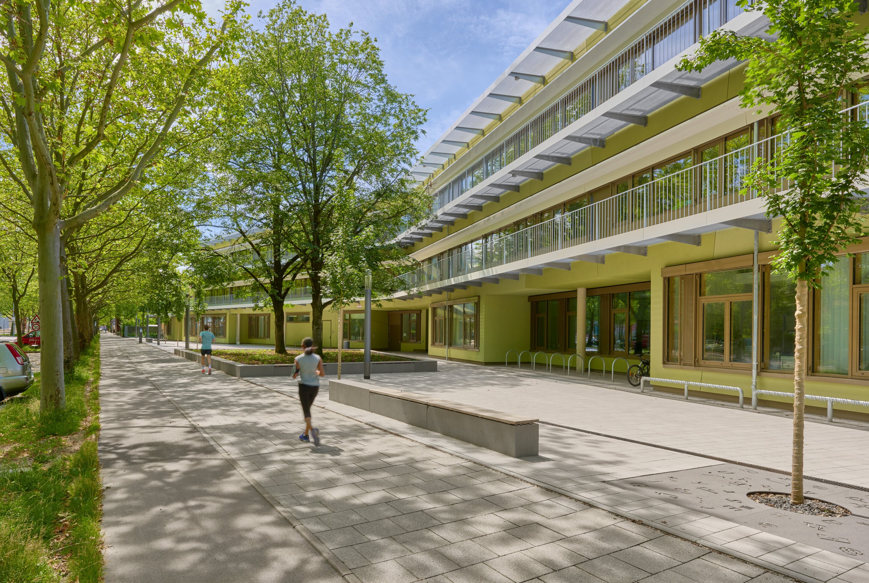 Project by Behnisch Architekturbuero, Primaryschool Infanteriestra&szlig;e. Tree-lined walkway beside a modern building with balconies, where people are walking and jogging along the paved path.
