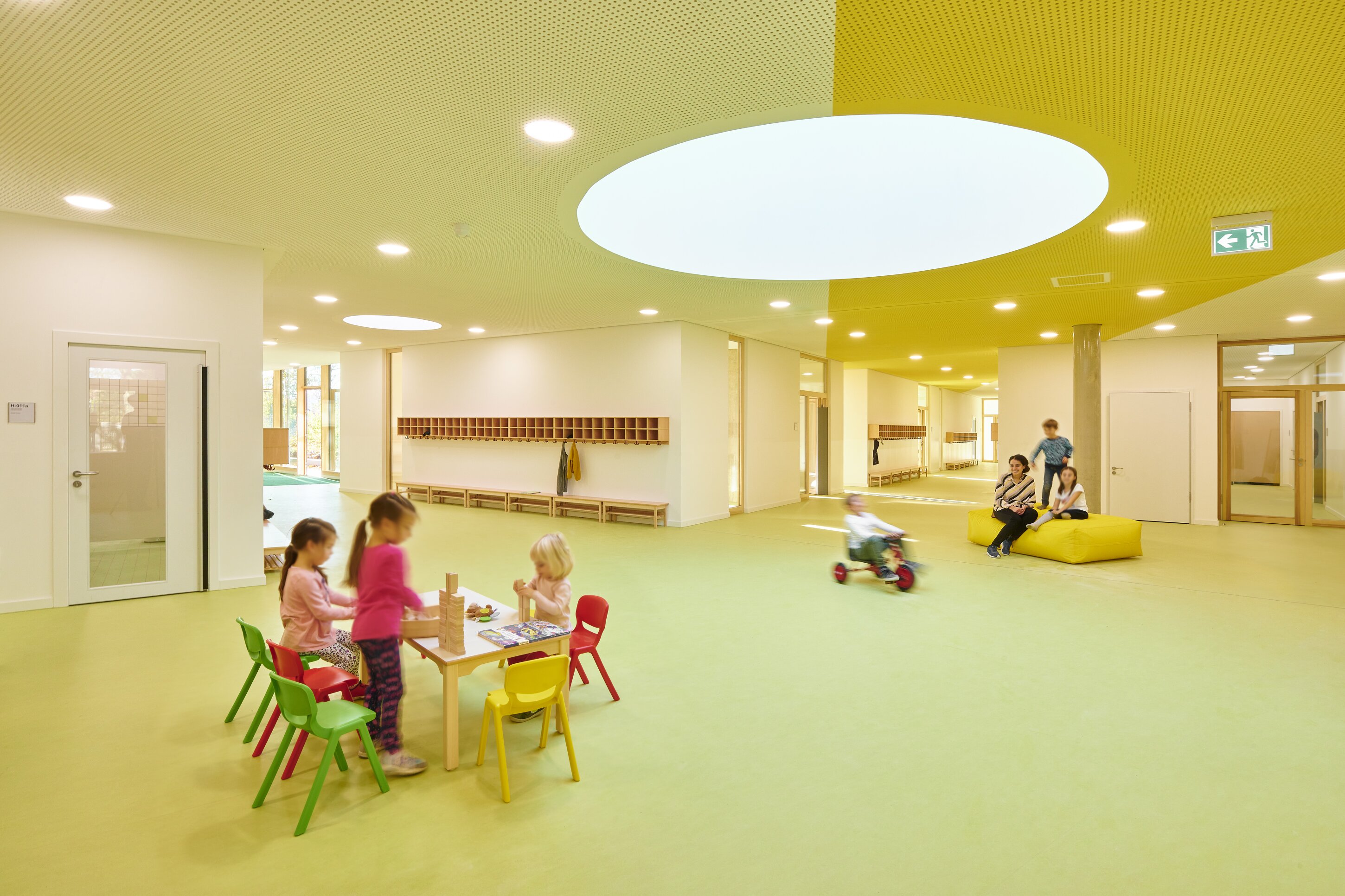 Project by Behnisch Architekturbuero, Primaryschool Infanteriestra&szlig;e. Bright indoor play area with children at a small table, others riding and sitting, colorful chairs, and a yellow ceiling with circular skylights.