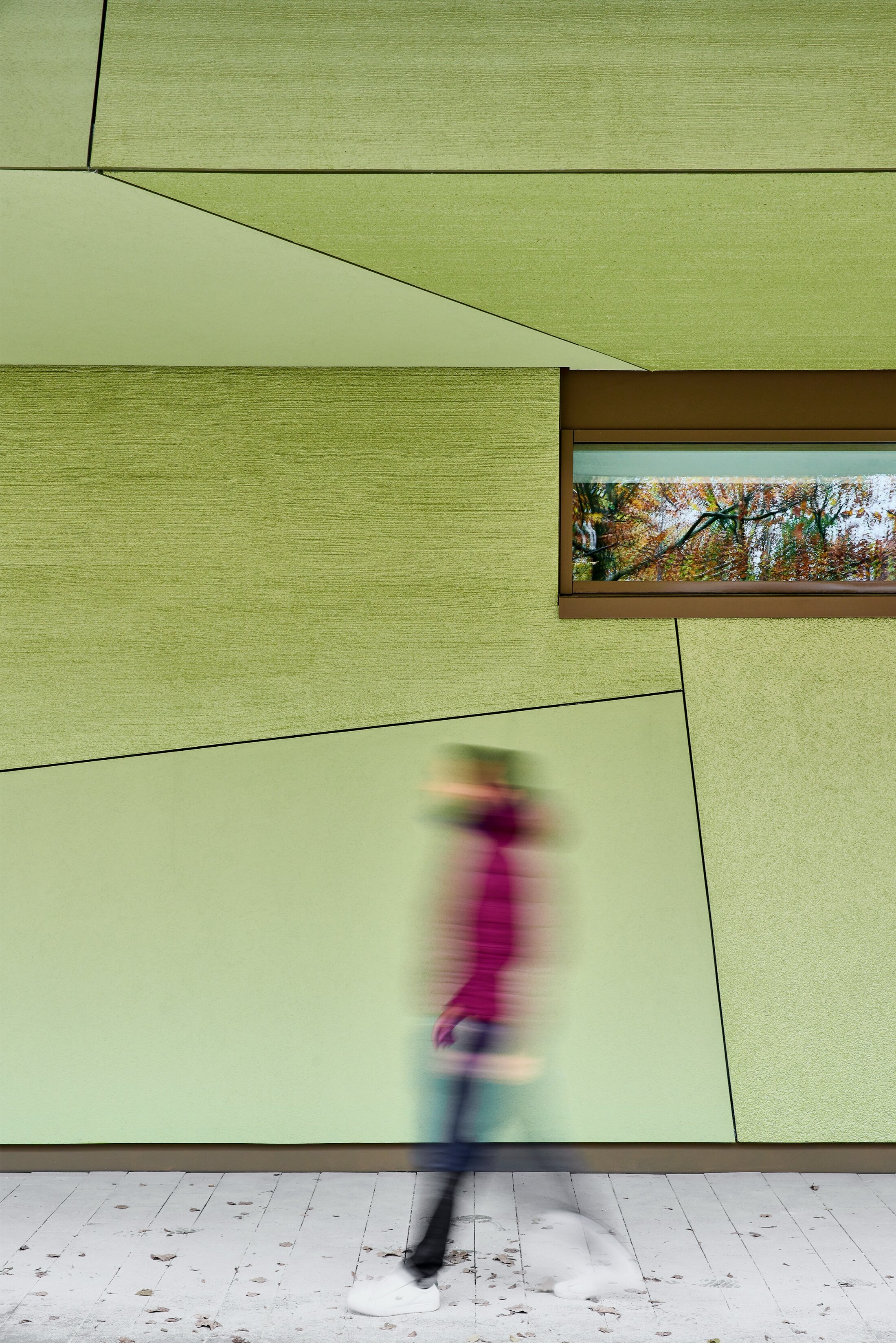 Project by Behnisch Architekturbuero, Primaryschool Infanteriestra&szlig;e. Green geometric building facade with a narrow window, and a blurred person walking past in the foreground.