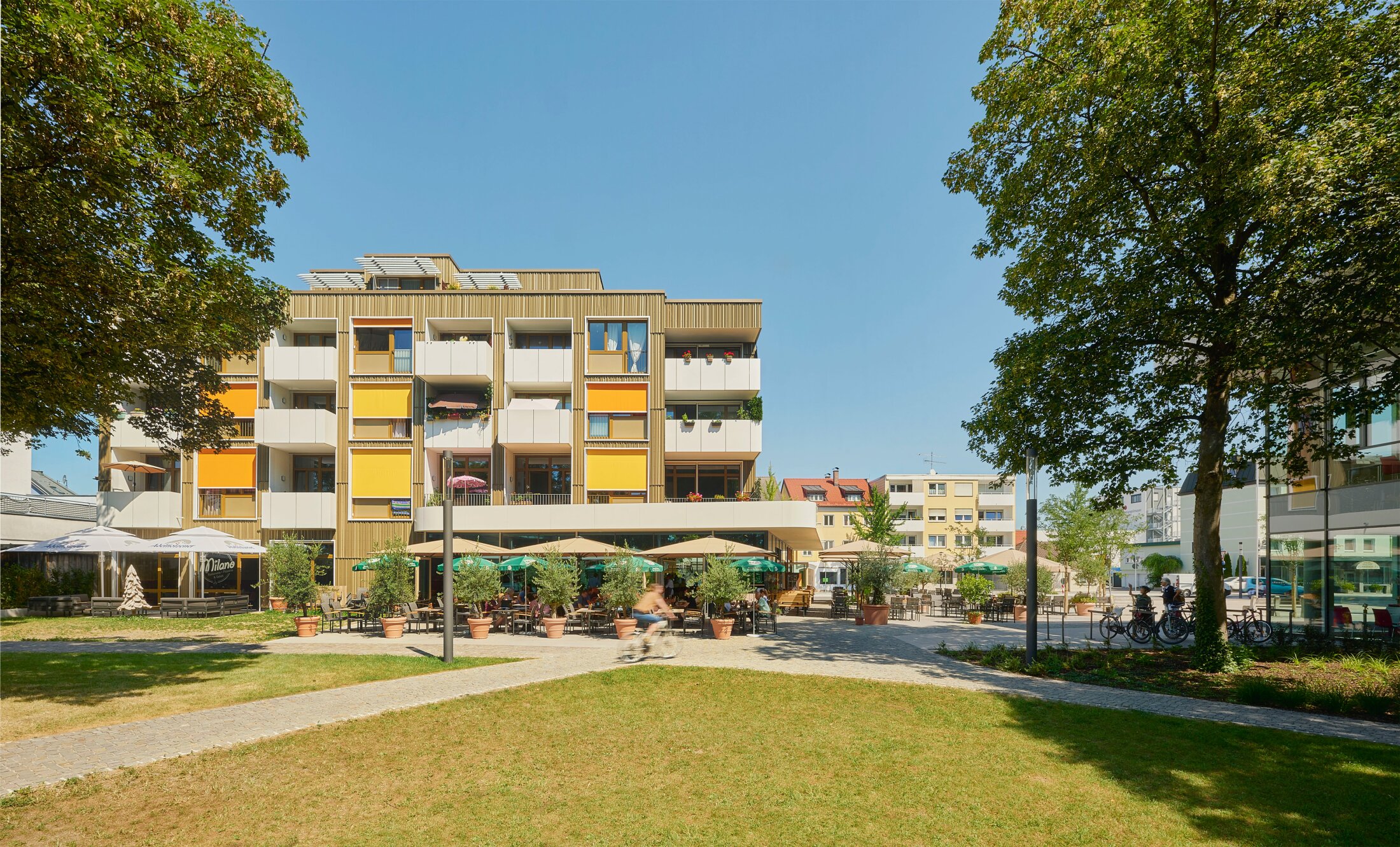 Project by Behnisch Architekturbuero, Mixed-use Building Rathausplatz. Apartment building with balconies, orange and yellow shades, caf&eacute; terrace below, trees and lawn in foreground, and people cycling past.
