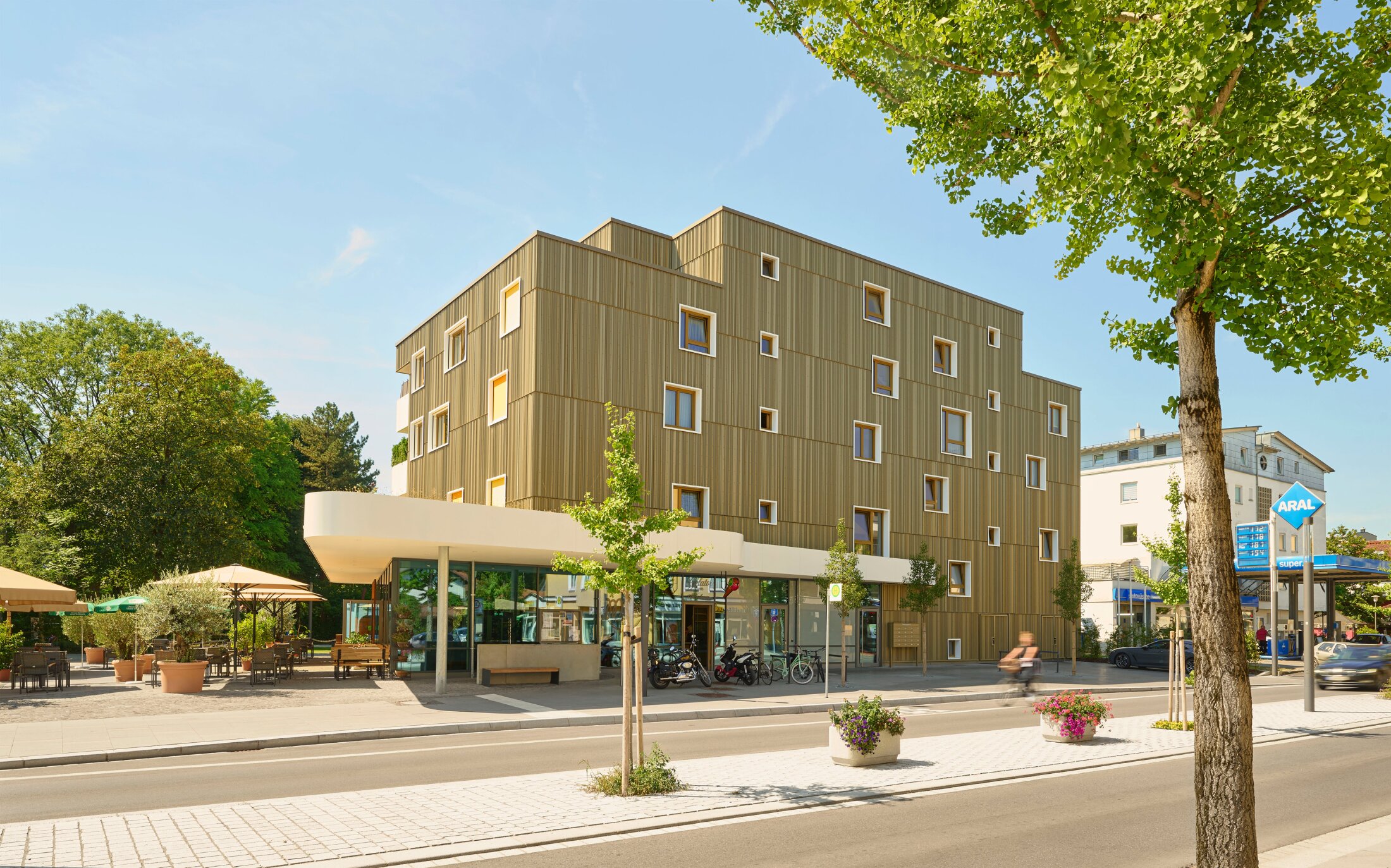 Project by Behnisch Architekturbuero, Mixed-use Building Rathausplatz. Modern wood-clad building with small square windows, street and caf&eacute; seating in front, trees and cyclist passing by.