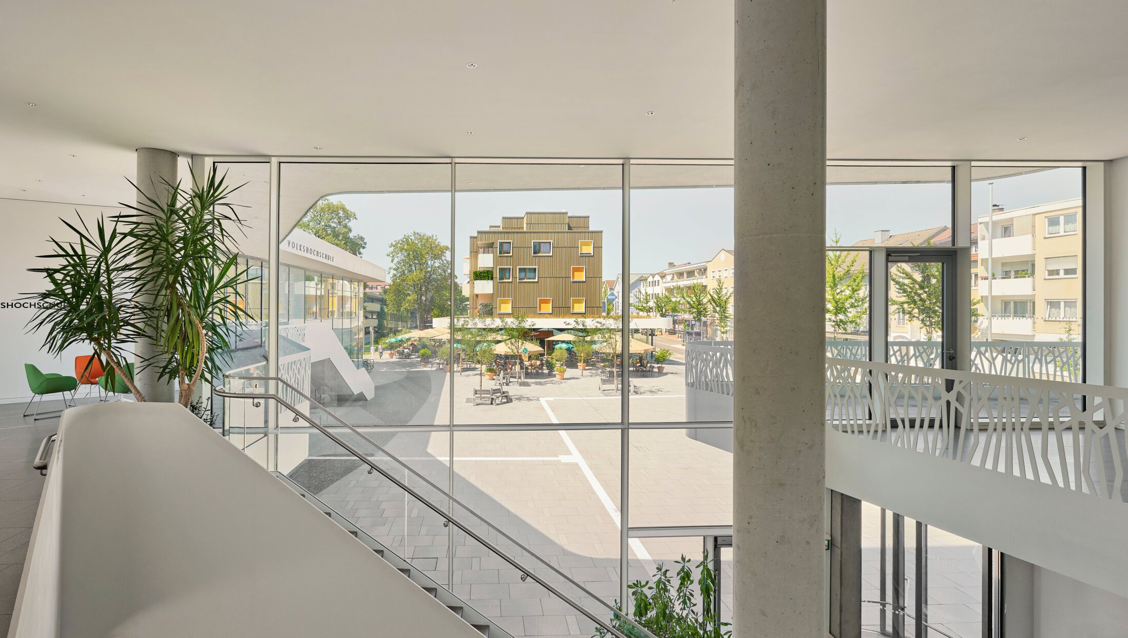 Project by Behnisch Architekturbuero, Mixed-use Building Rathausplatz. Interior view with staircase and large windows overlooking the plaza with caf&eacute; seating and a modern wood-clad building outside.