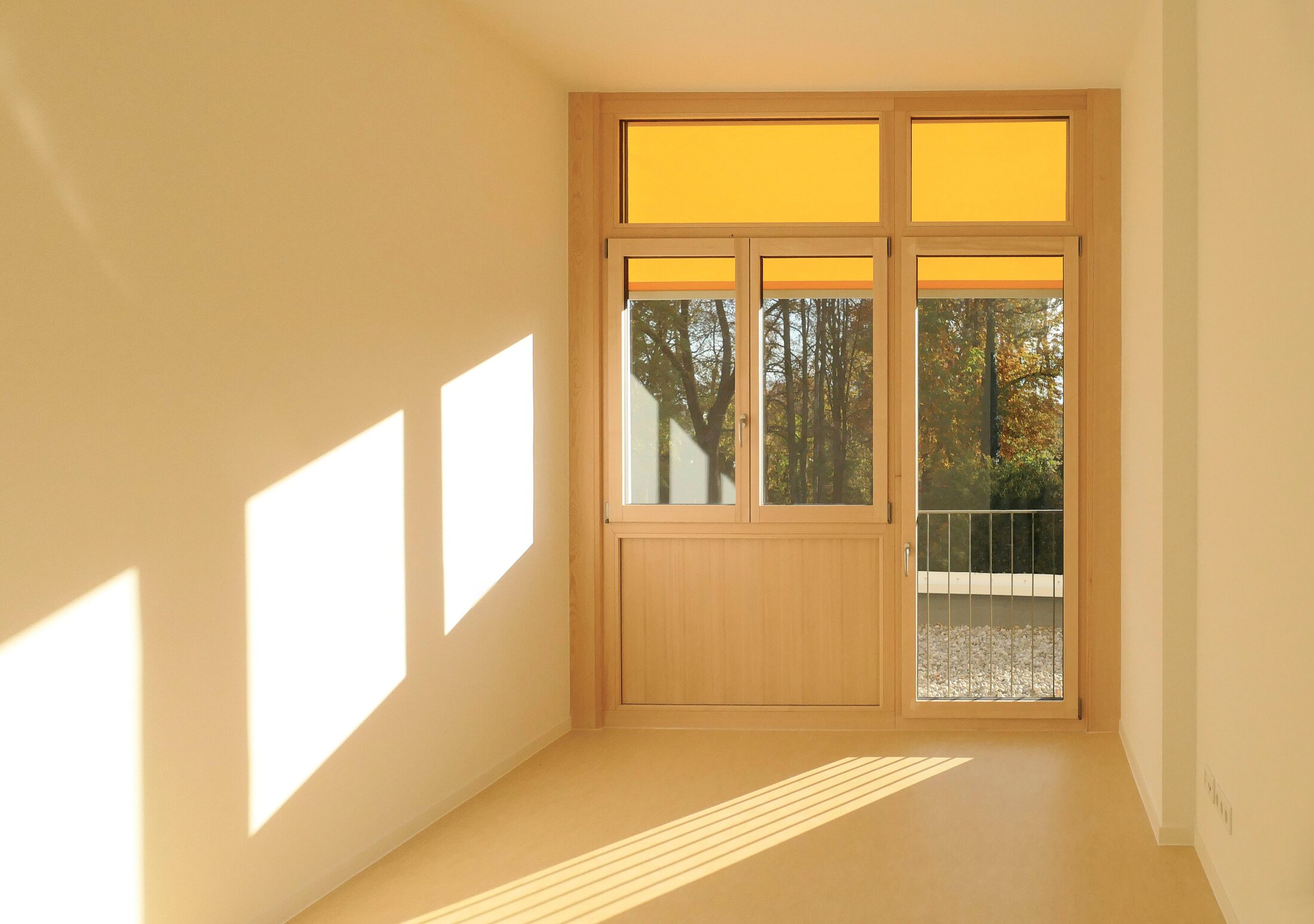 Project by Behnisch Architekturbuero, Mixed-use Building Rathausplatz. Room with light wood-framed window and door, warm sunlight casting rectangular shadows on white walls, trees visible outside.