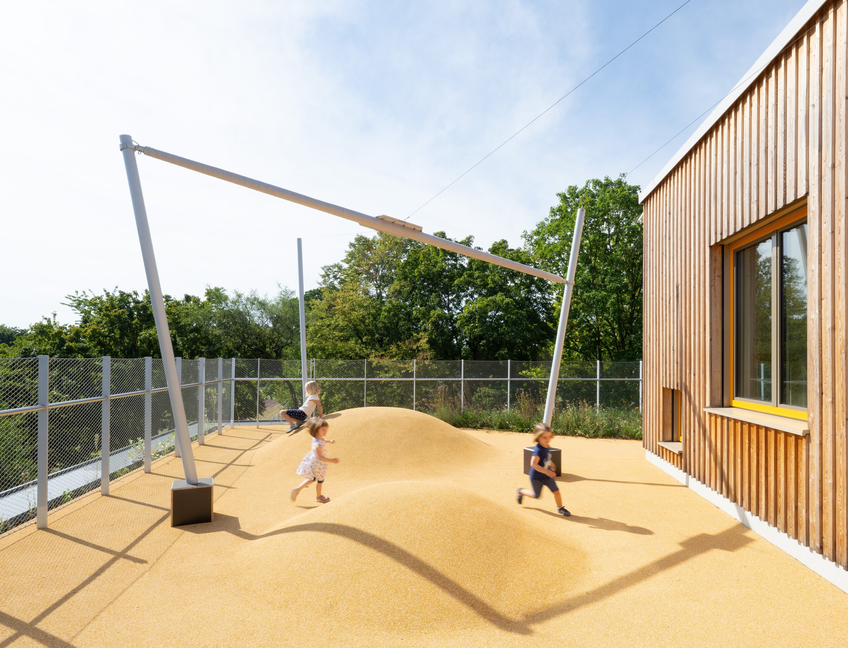 Project by Behnisch Architekturbuero, Family Center R&ouml;thelheim 11acht and Gerd Lohwasser Sports Hall. Children playing on a small mound playground beside a wooden building, with a metal frame structure and fenced area under a clear sky.