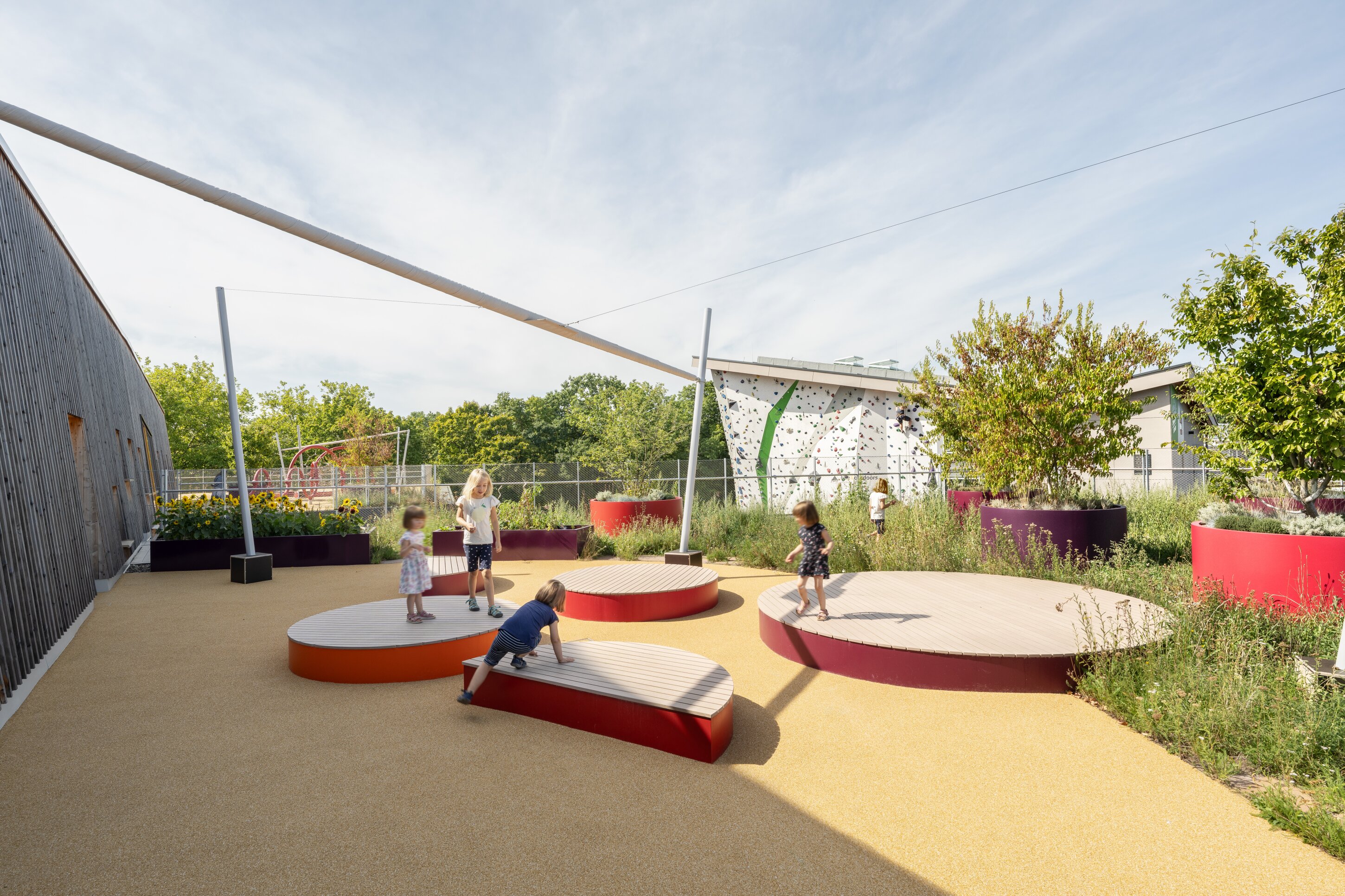Project by Behnisch Architekturbuero, Family Center R&ouml;thelheim 11acht and Gerd Lohwasser Sports Hall. Children playing on round platforms in a playground courtyard, with shade canopy, planting beds, and a climbing wall in the background.