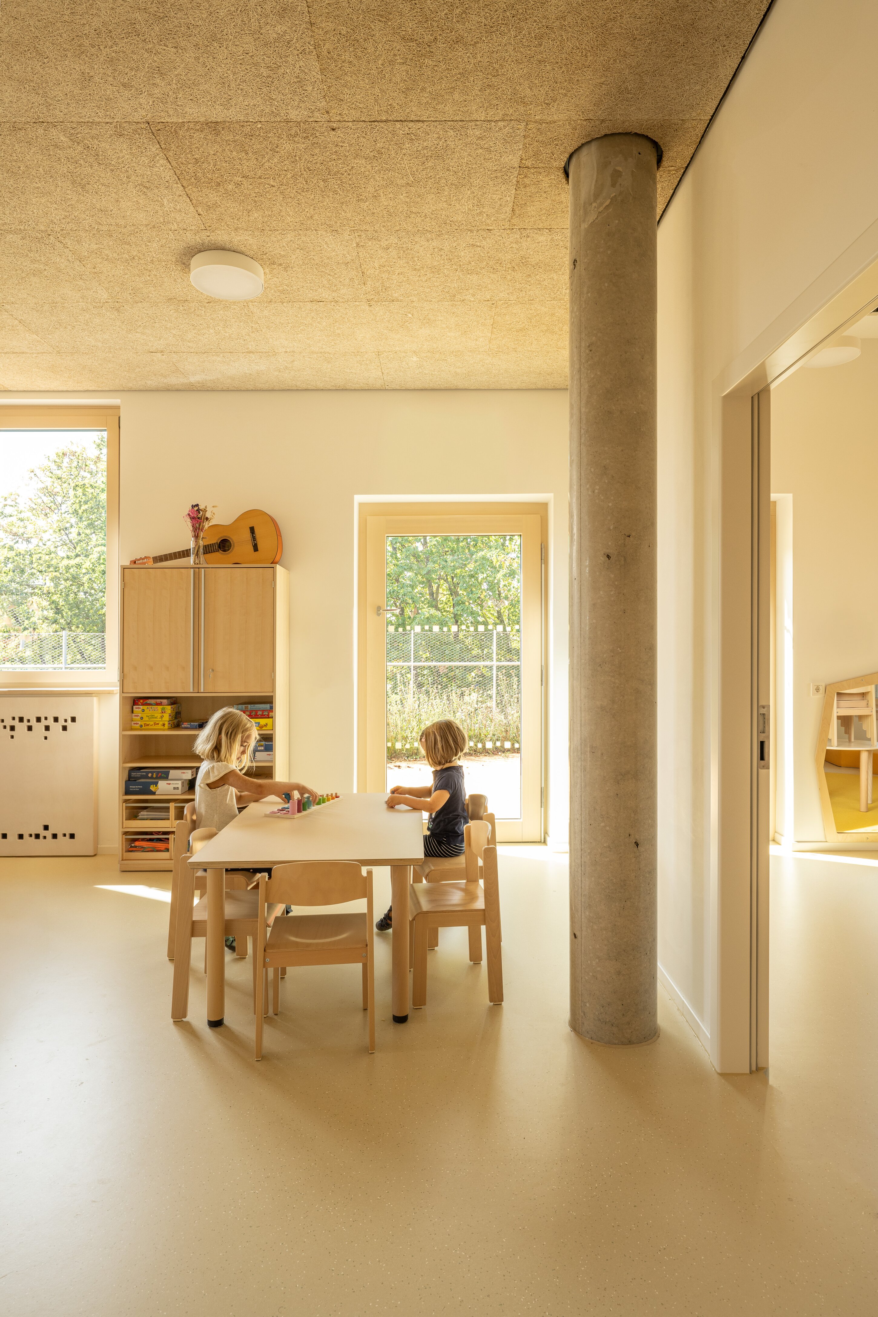 Project by Behnisch Architekturbuero, Family Center R&ouml;thelheim 11acht and Gerd Lohwasser Sports Hall. Two children sit at a small table in a bright classroom, playing with toys; wooden shelves, a guitar, and a large window are in the background.
