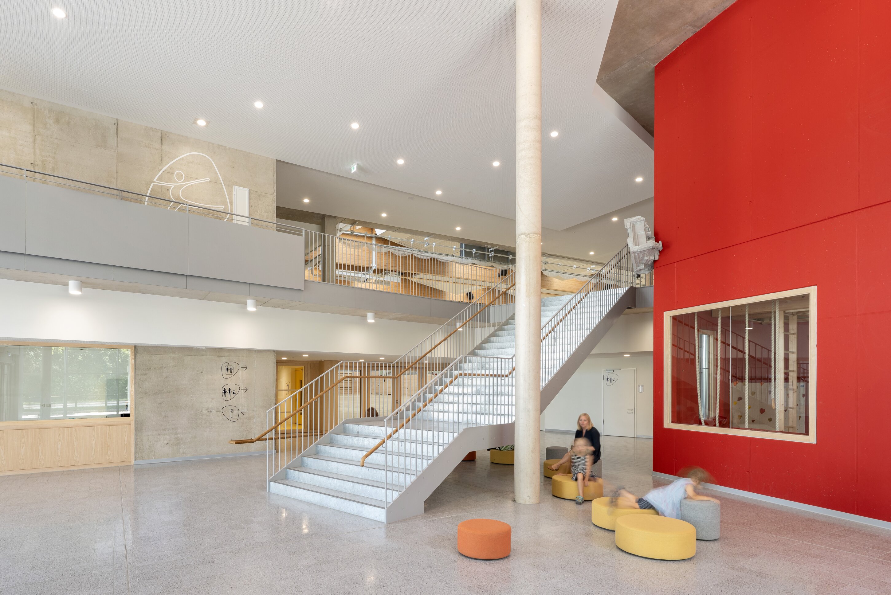Project by Behnisch Architekturbuero, Family Center R&ouml;thelheim 11acht and Gerd Lohwasser Sports Hall. Bright interior atrium with a central staircase, concrete column, red accent wall, and children playing on round seats near an adult.