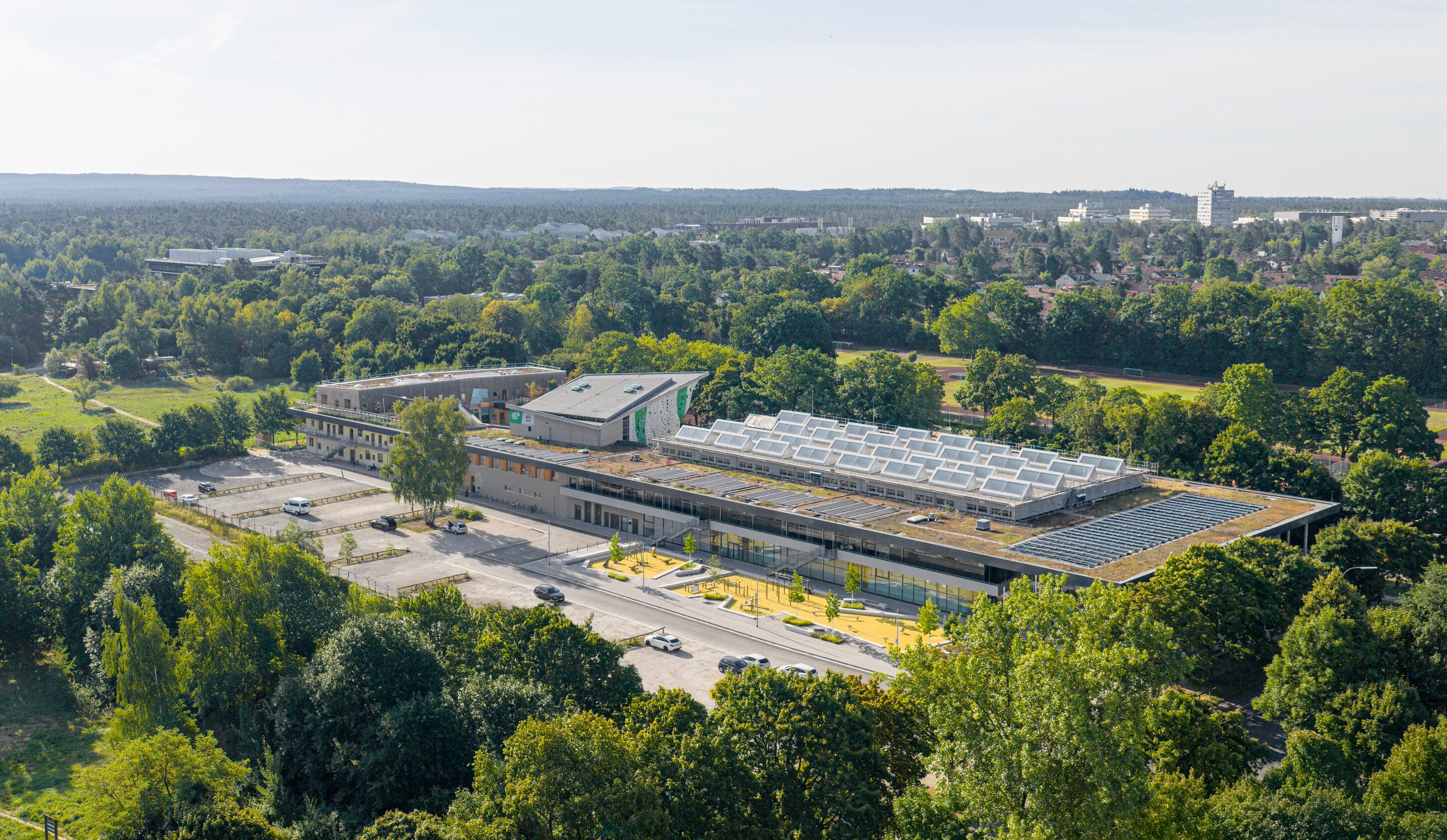 Project by Behnisch Architekturbuero, Family Center R&ouml;thelheim 11acht and Gerd Lohwasser Sports Hall. Aerial view of the campus building with green roofs and skylights, surrounded by trees, parking areas, and nearby residential neighborhood.