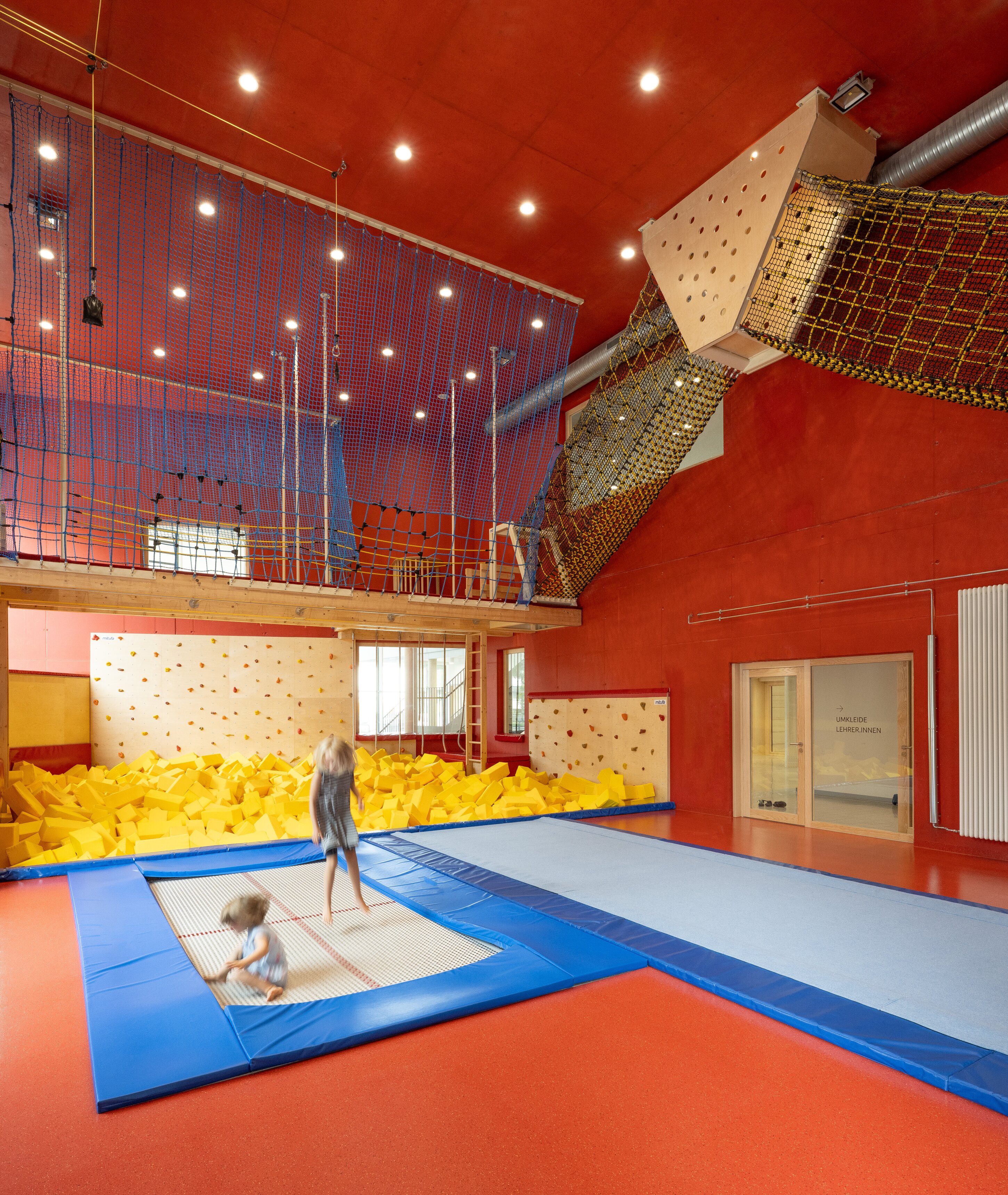 Project by Behnisch Architekturbuero, Family Center R&ouml;thelheim 11acht and Gerd Lohwasser Sports Hall. Indoor gym play area with trampolines, foam pit, climbing wall, and netted play structure; two children jumping on a trampoline.