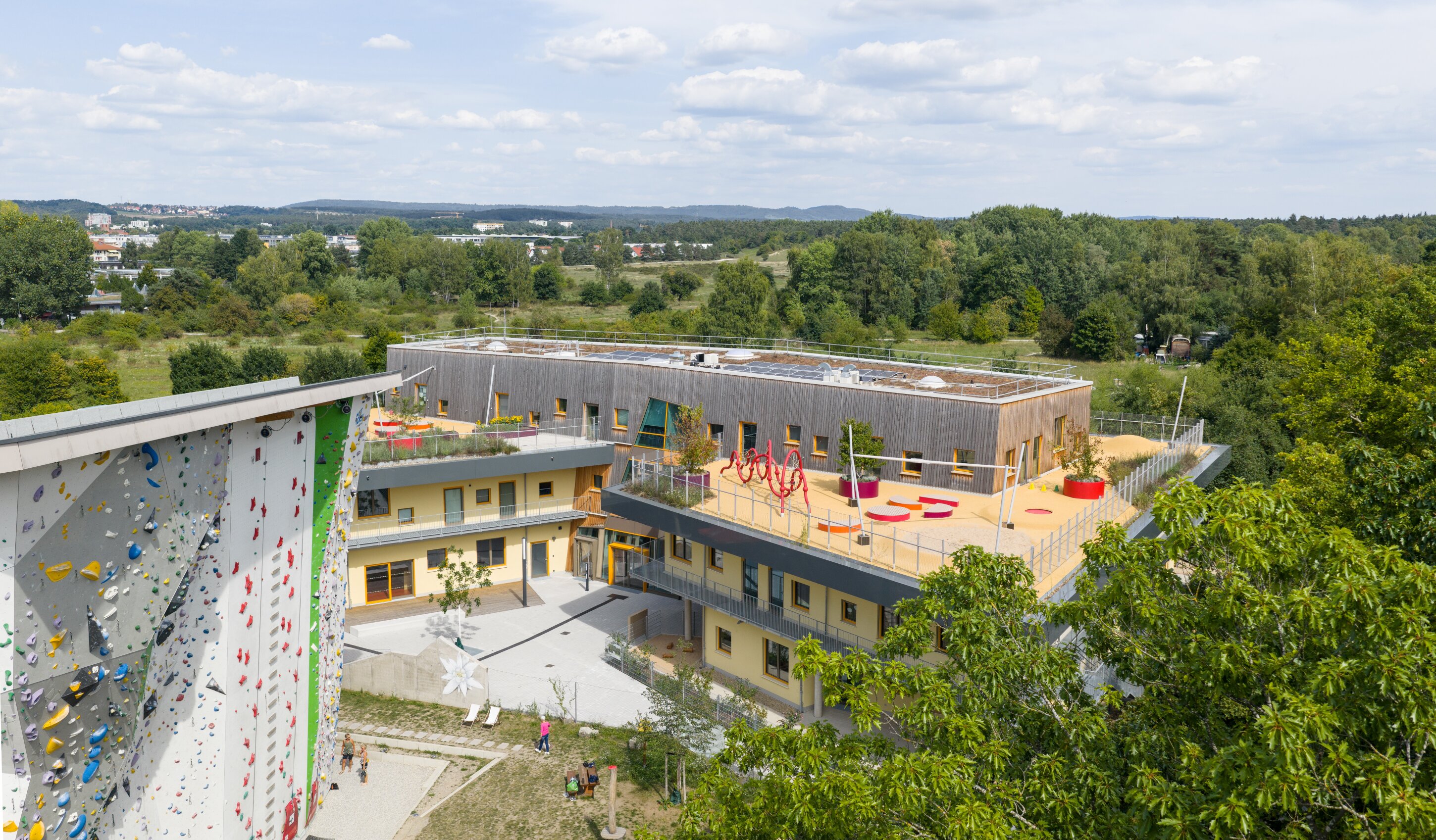 Project by Behnisch Architekturbuero, Family Center R&ouml;thelheim 11acht and Gerd Lohwasser Sports Hall. Aerial view of a school complex with a rooftop playground, courtyard, and adjacent climbing wall, surrounded by trees and open landscape.