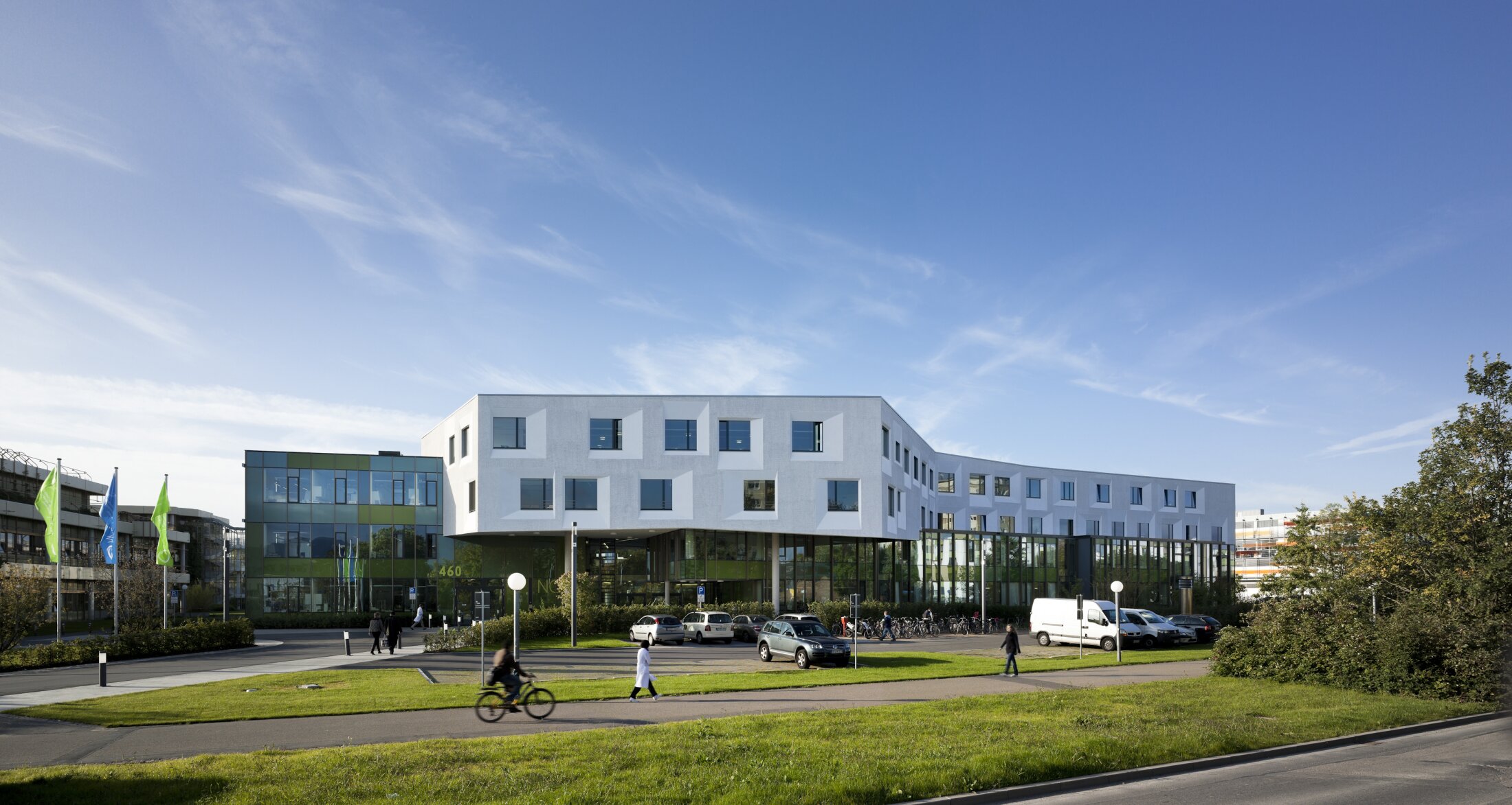 Project by Behnisch Architekturbuero, NCT National Center for Tumour Diseases. Modern building with white and glass facades, surrounded by grass, parked cars, and pedestrians under a blue sky.