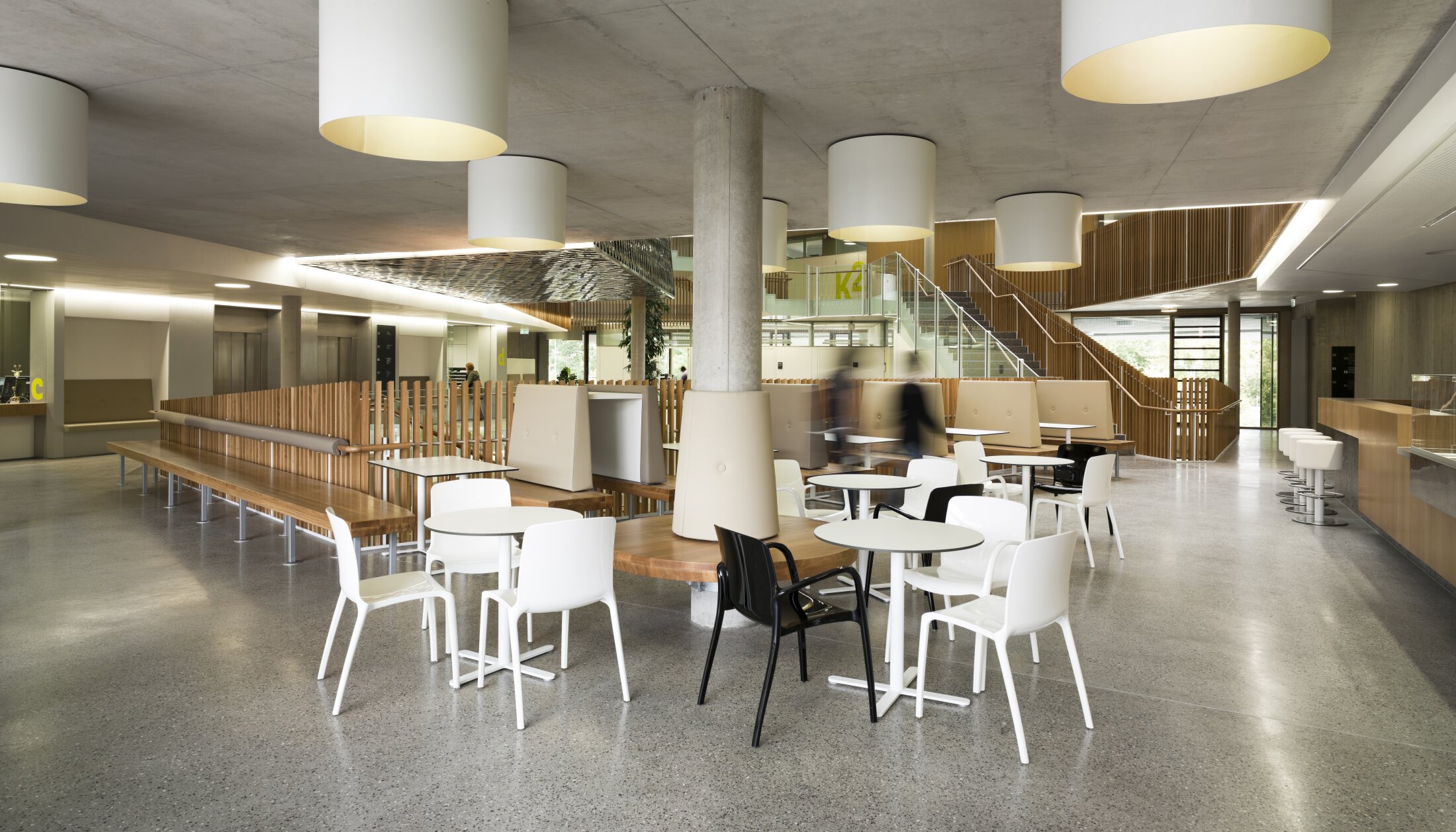 Project by Behnisch Architekturbuero, NCT National Center for Tumour Diseases. Open cafeteria with tables and chairs, wooden benches, large ceiling lights, stairs and people moving around in the modern common room.