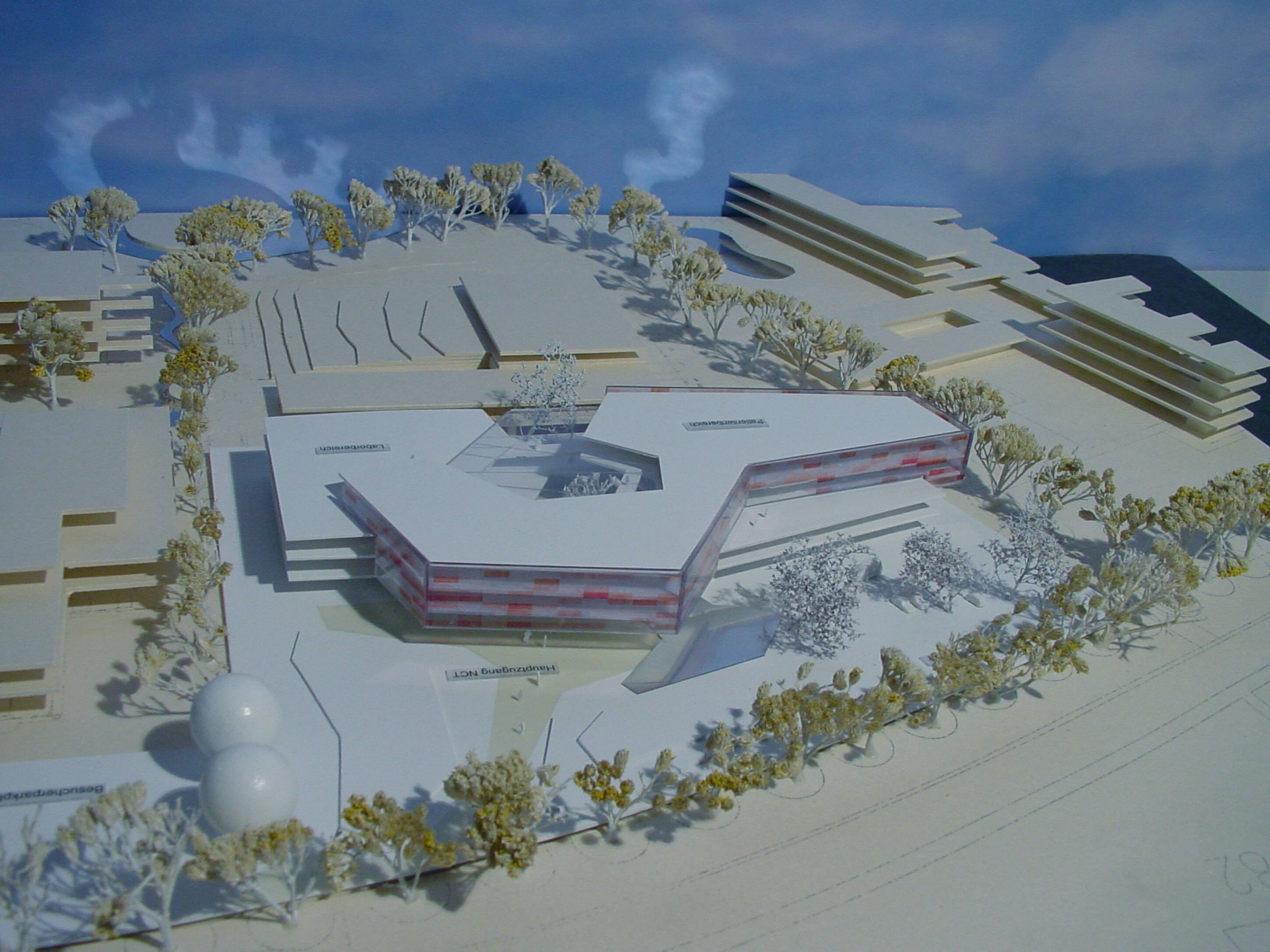 Project by Behnisch Architekturbuero, NCT National Center for Tumour Diseases. Architectural site model showing a low, angular building with a central courtyard, surrounded by trees and adjacent linear structures.