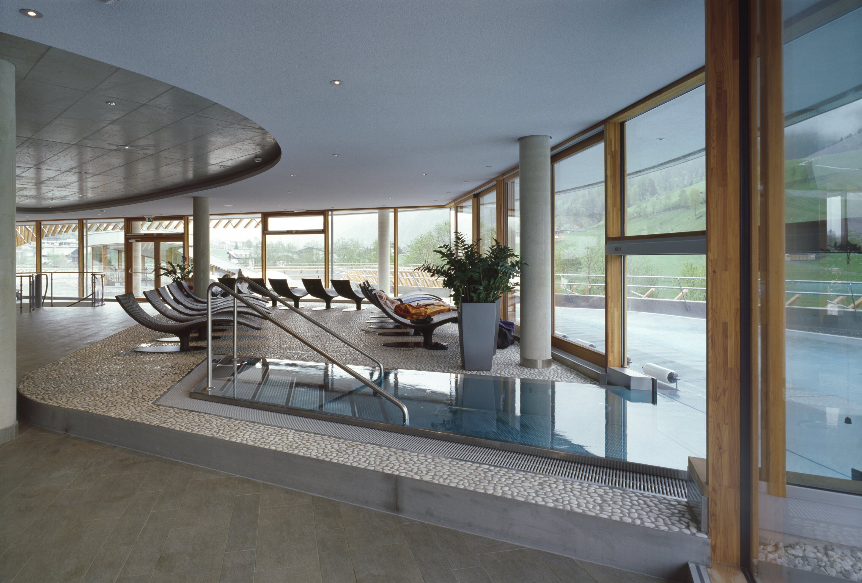 Project by Behnisch Architekturbuero, Thermal Roemerbad. Indoor spa area with pool, lounge chairs, and large windows overlooking a green hillside landscape.