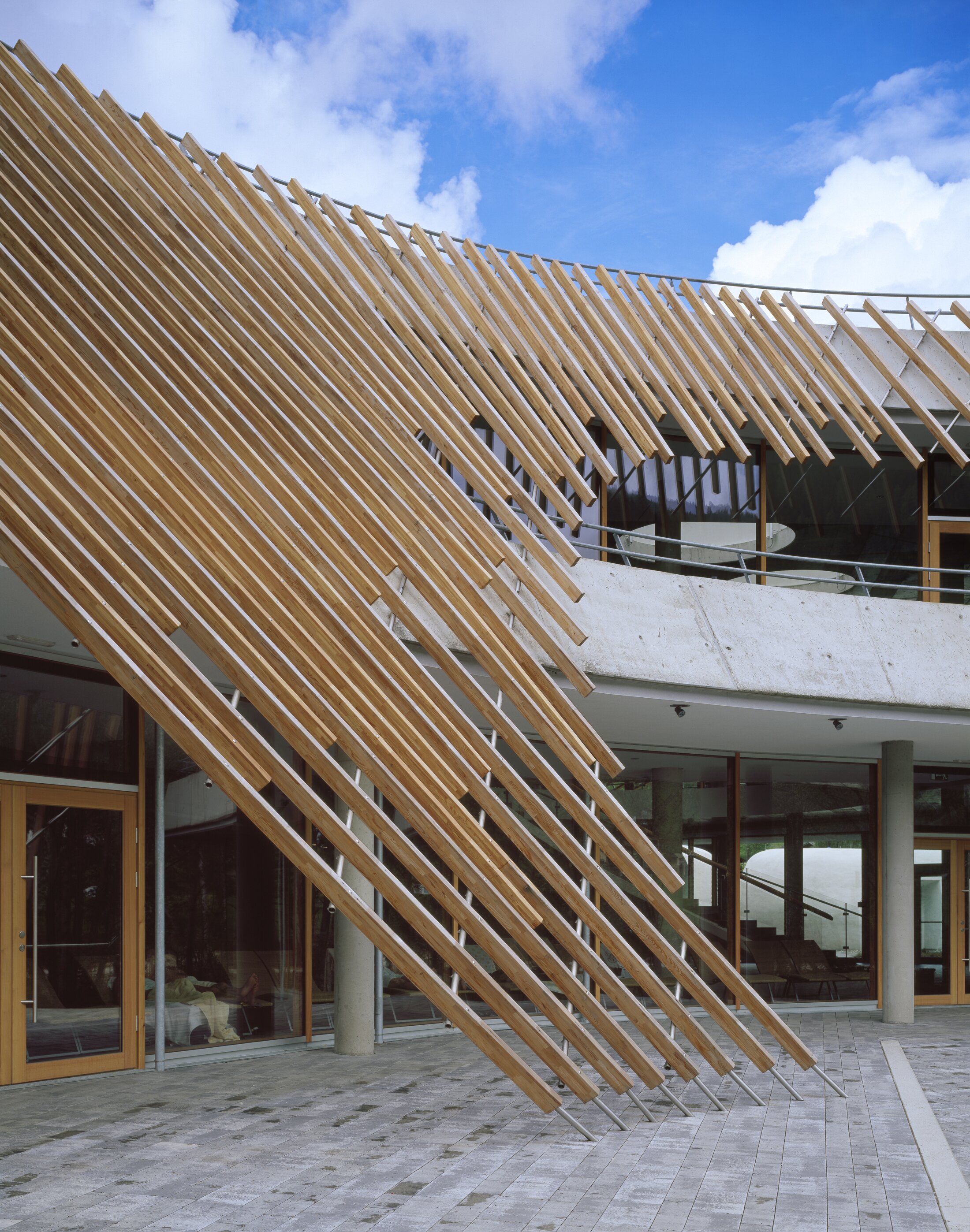 Project by Behnisch Architekturbuero, Thermal Roemerbad. Slanted wooden slats form a facade above the entrance of the glass building; above this is a curved concrete structure and blue sky. Through the glass facade, one can see the interior of the thermal baths with loungers and bathers.