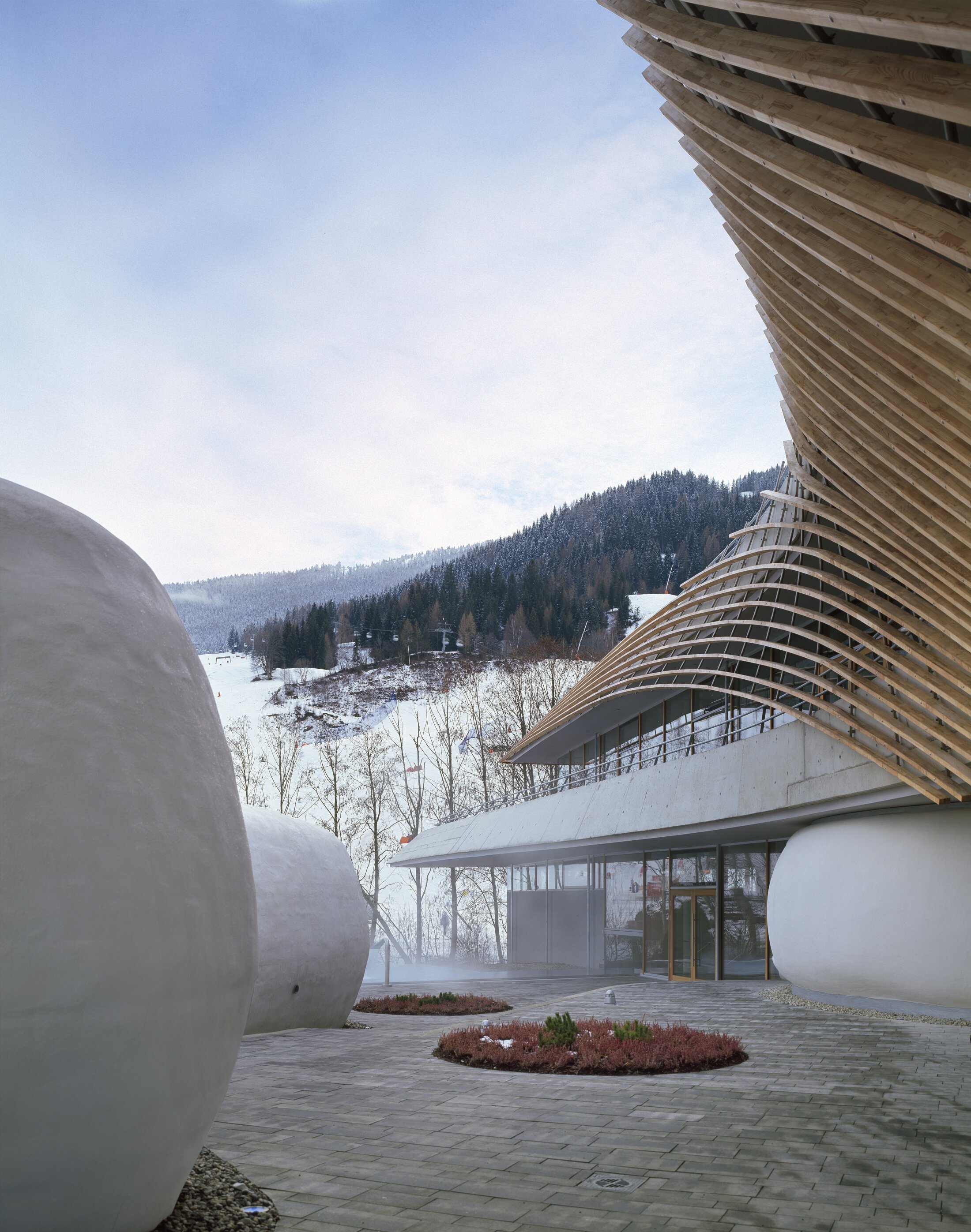 Project by Behnisch Architekturbuero, Thermal Roemerbad. Curved wooden roof over glass building with large rounded structures in foreground, set against snowy hillside and trees.