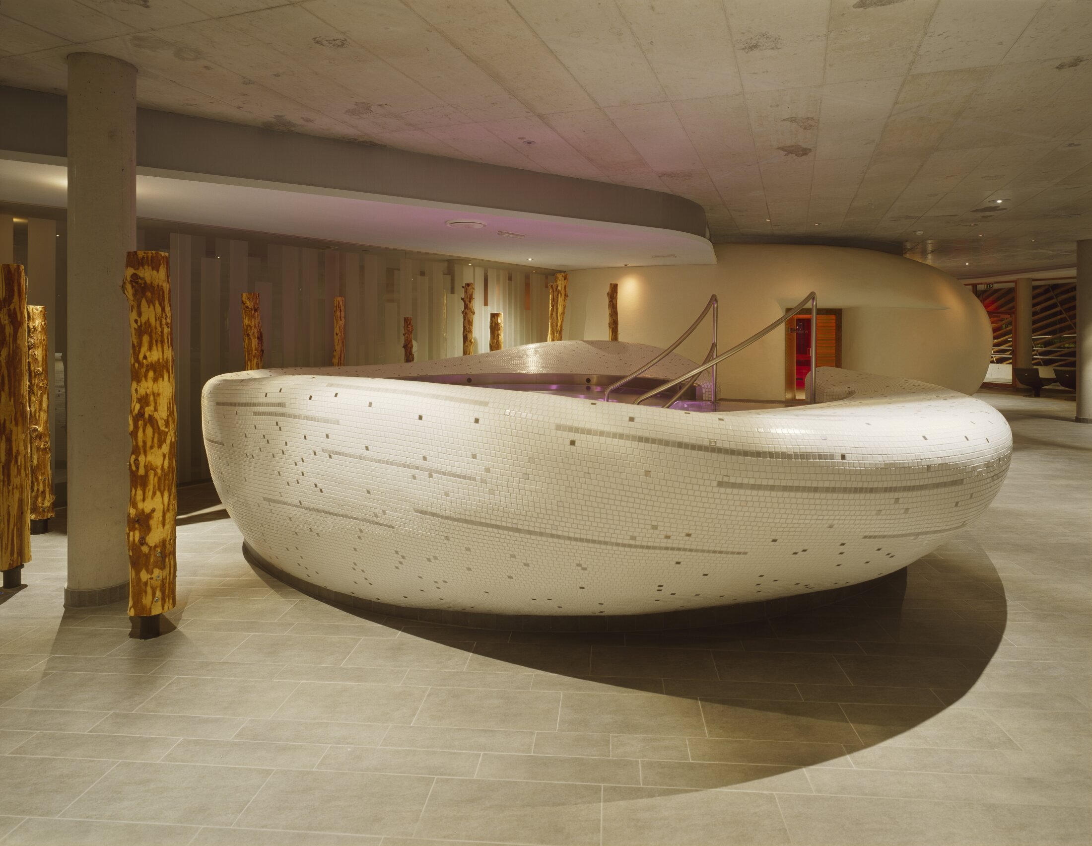 Project by Behnisch Architekturbuero, Thermal Roemerbad. Curved tiled hot tub in a spa interior with wooden columns, soft lighting, and a smooth concrete ceiling.