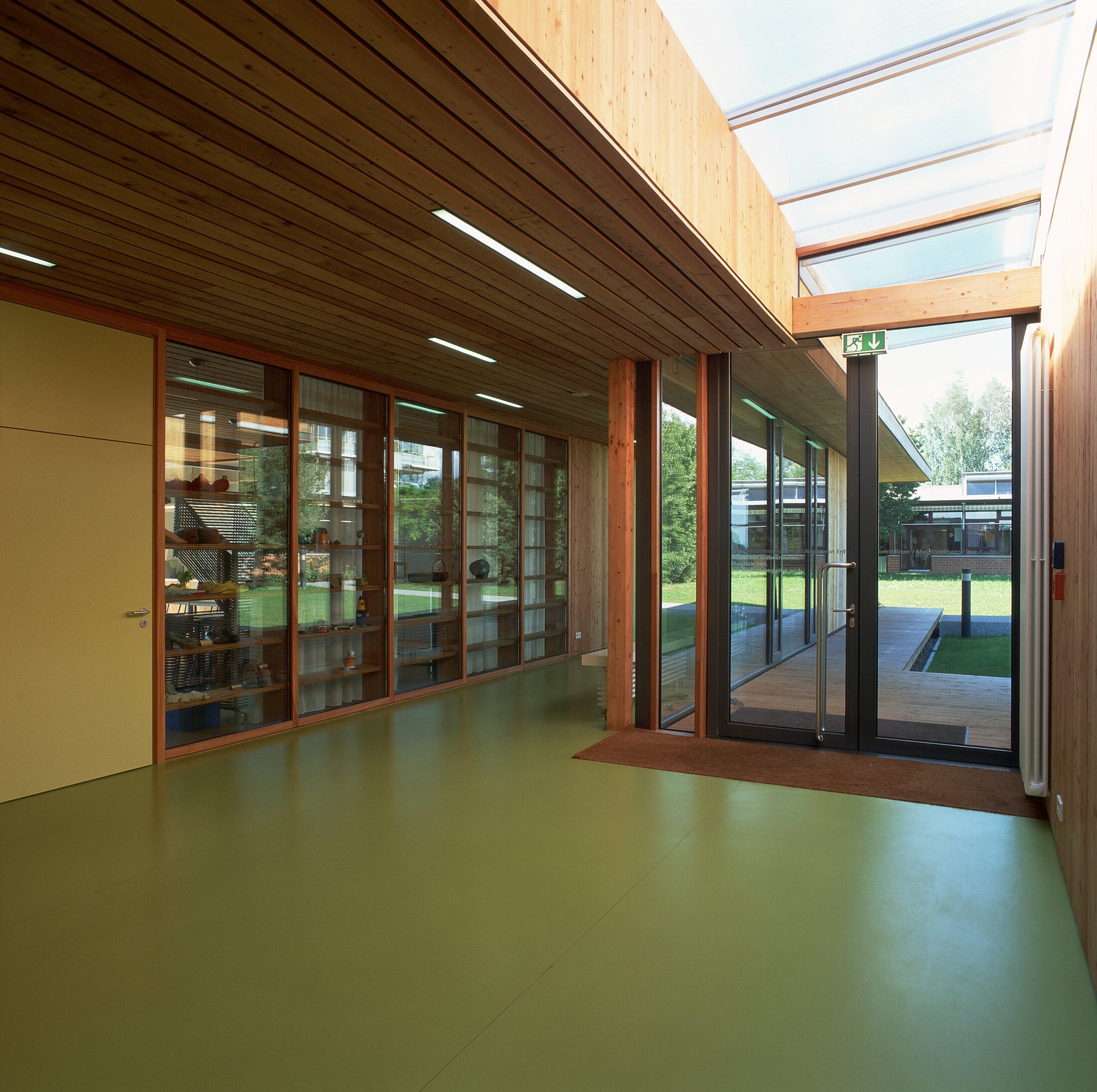 Project by Behnisch Architekturbuero, Hilde Domin School. A wood-lined hallway with a green floor, glass partitions and glass entrance doors that open onto a courtyard illuminated by skylights.
