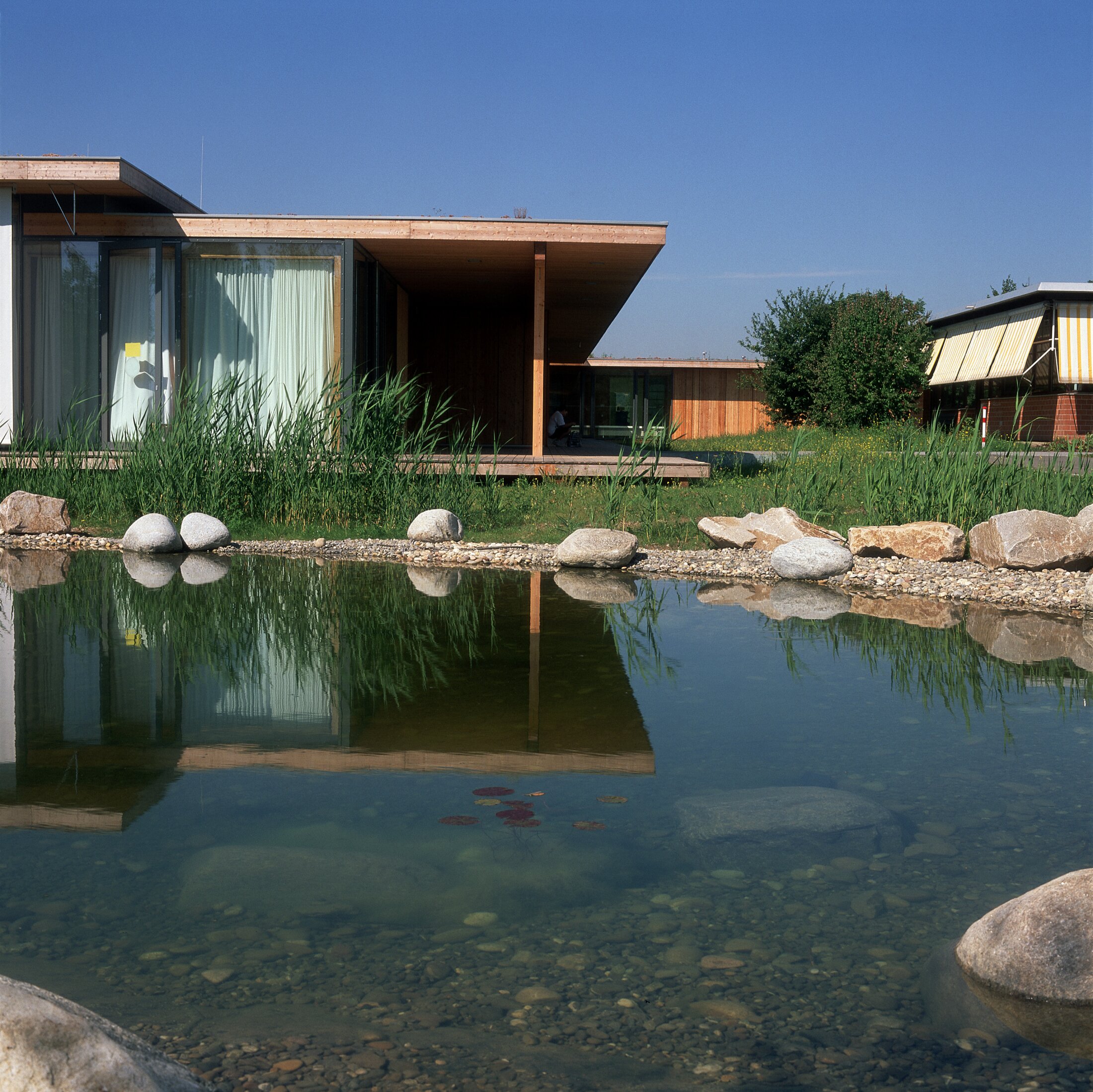 Project by Behnisch Architekturbuero, Hilde Domin School. A modern wooden house stands beside a pond, its waters reflecting rocks and reeds. The large, floor-to-ceiling windows are darkened with curtains. The right side of the building is also visible and features external shading.