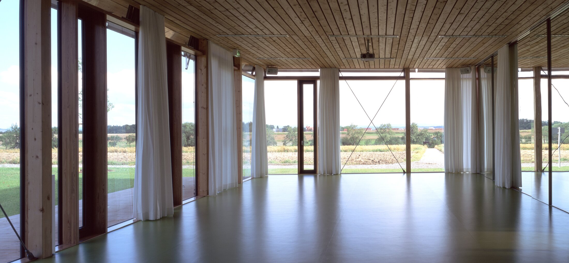 Project by Behnisch Architekturbuero, Hilde Domin School. Empty room with wooden ceiling, green floor, and floor-to-ceiling windows with curtains overlooking fields and trees.