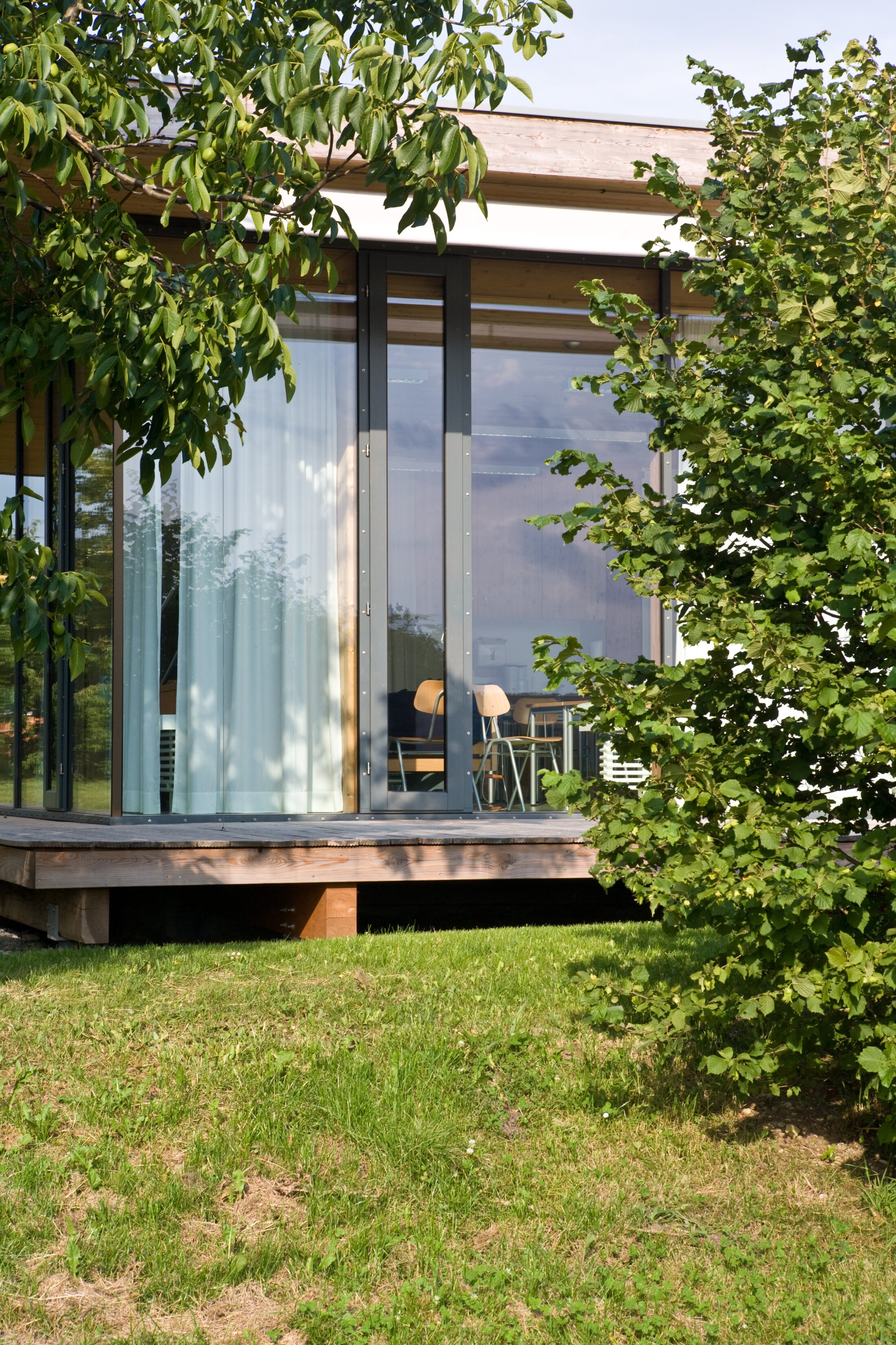 Project by Behnisch Architekturbuero, Hilde Domin School. Exterior view of the building with a floor-to-ceiling glass facade that can be shaded with curtains. The elevated building is visible through leafy trees and sits on a lawn with a wooden terrace.