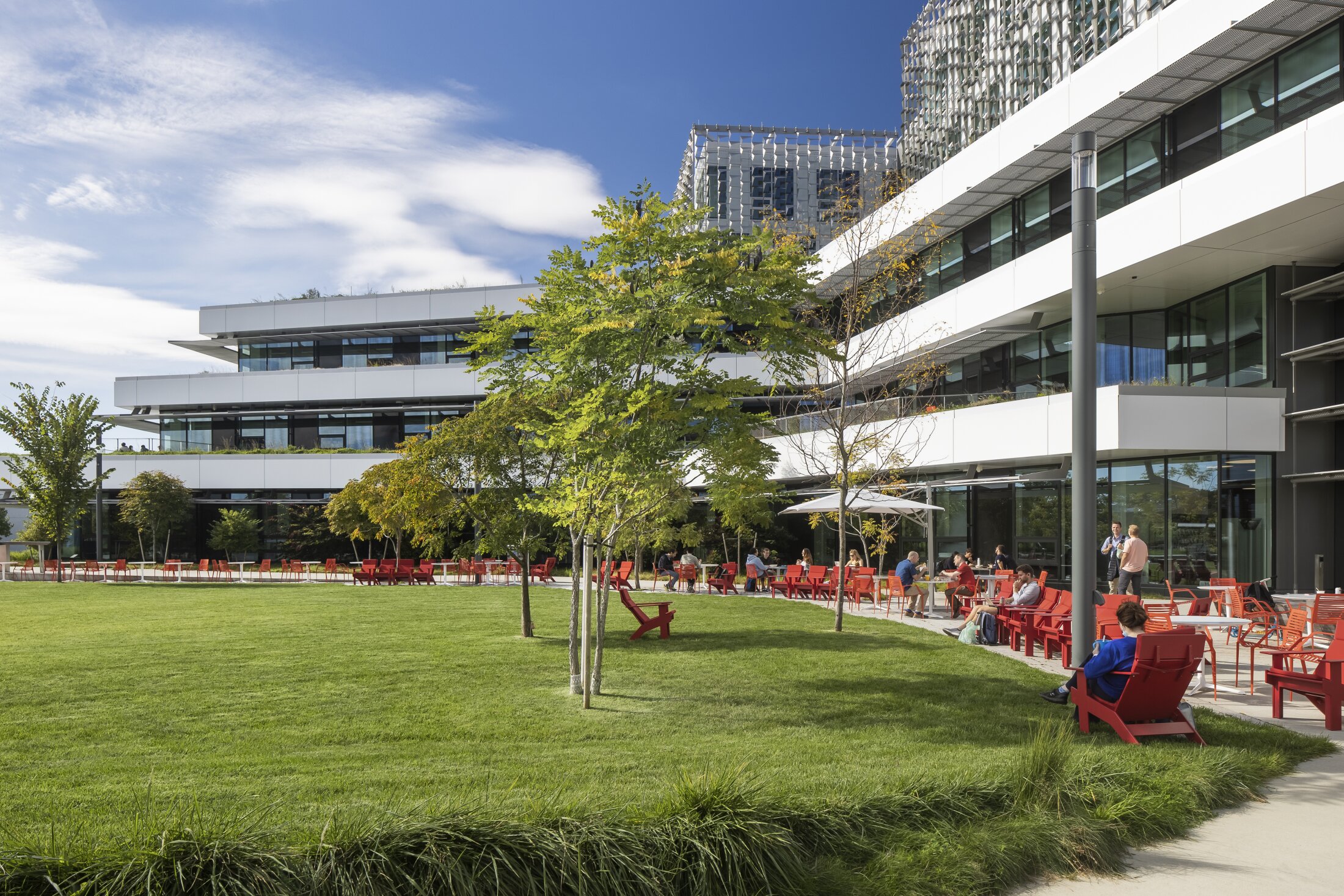 Project by Behnisch Architekturbuero, Harvard University Science and Engineering Complex (SEC). Modern office campus with glass buildings, lawn, trees, and people seated on red chairs along a terrace and outdoor seating area.