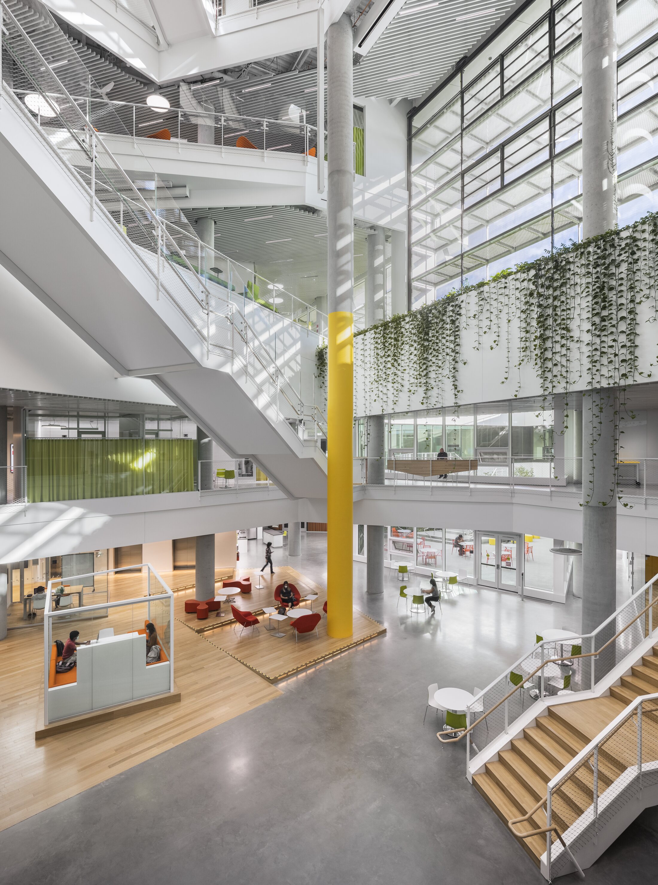 Project by Behnisch Architekturbuero, Harvard University Science and Engineering Complex (SEC). Bright multi-level atrium with stairs, glass walls, a tall yellow column, hanging plants, and seating areas below.