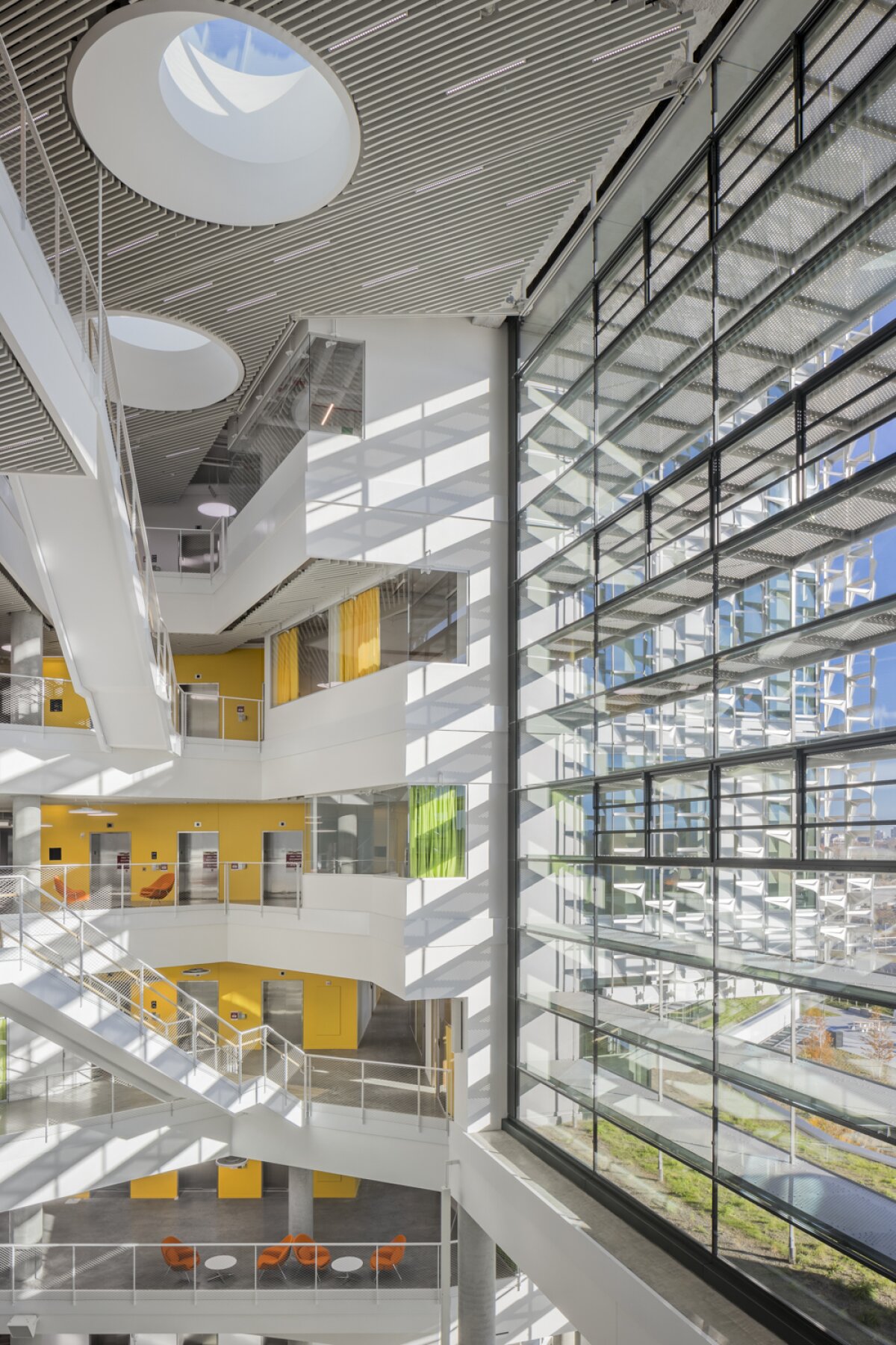 Project by Behnisch Architekturbuero, Harvard University Science and Engineering Complex (SEC). A bright, multi-story atrium with a glass facade, stairwells, balconies, and yellow accents, illuminated by daylight through large windows. Round skylights also provide additional light.
