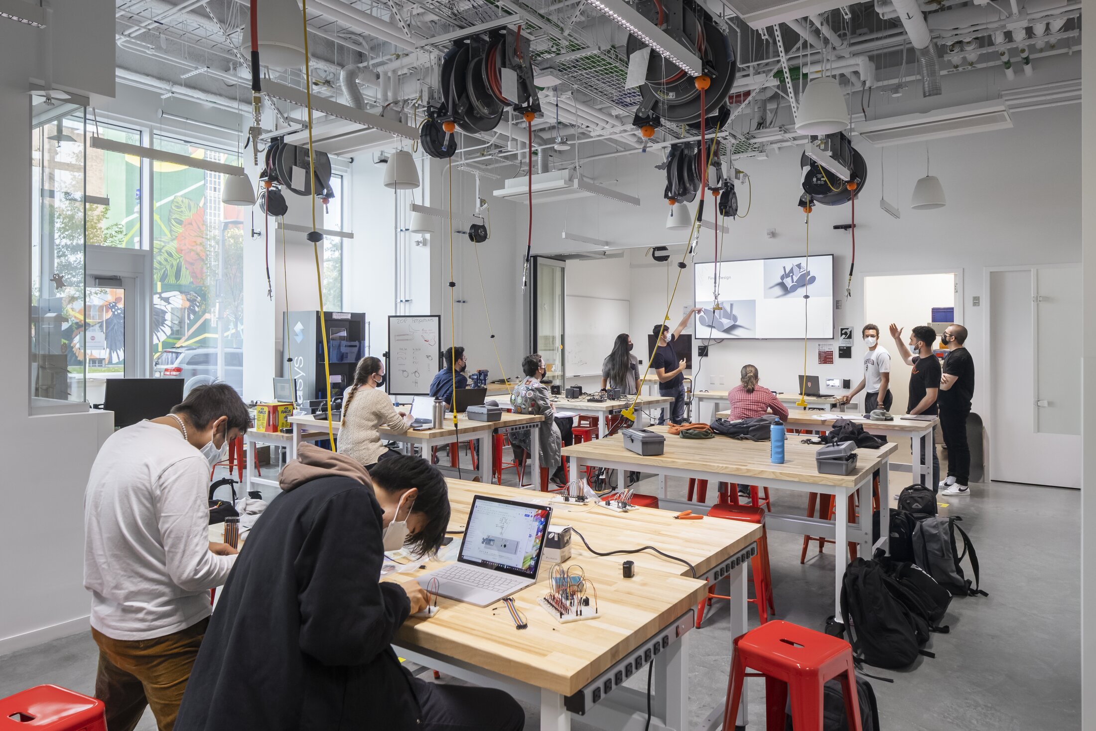 Project by Behnisch Architekturbuero, Harvard University Science and Engineering Complex (SEC). Students working at tables with laptops and electronics while an instructor and some students presents on a screen in a bright workshop classroom.