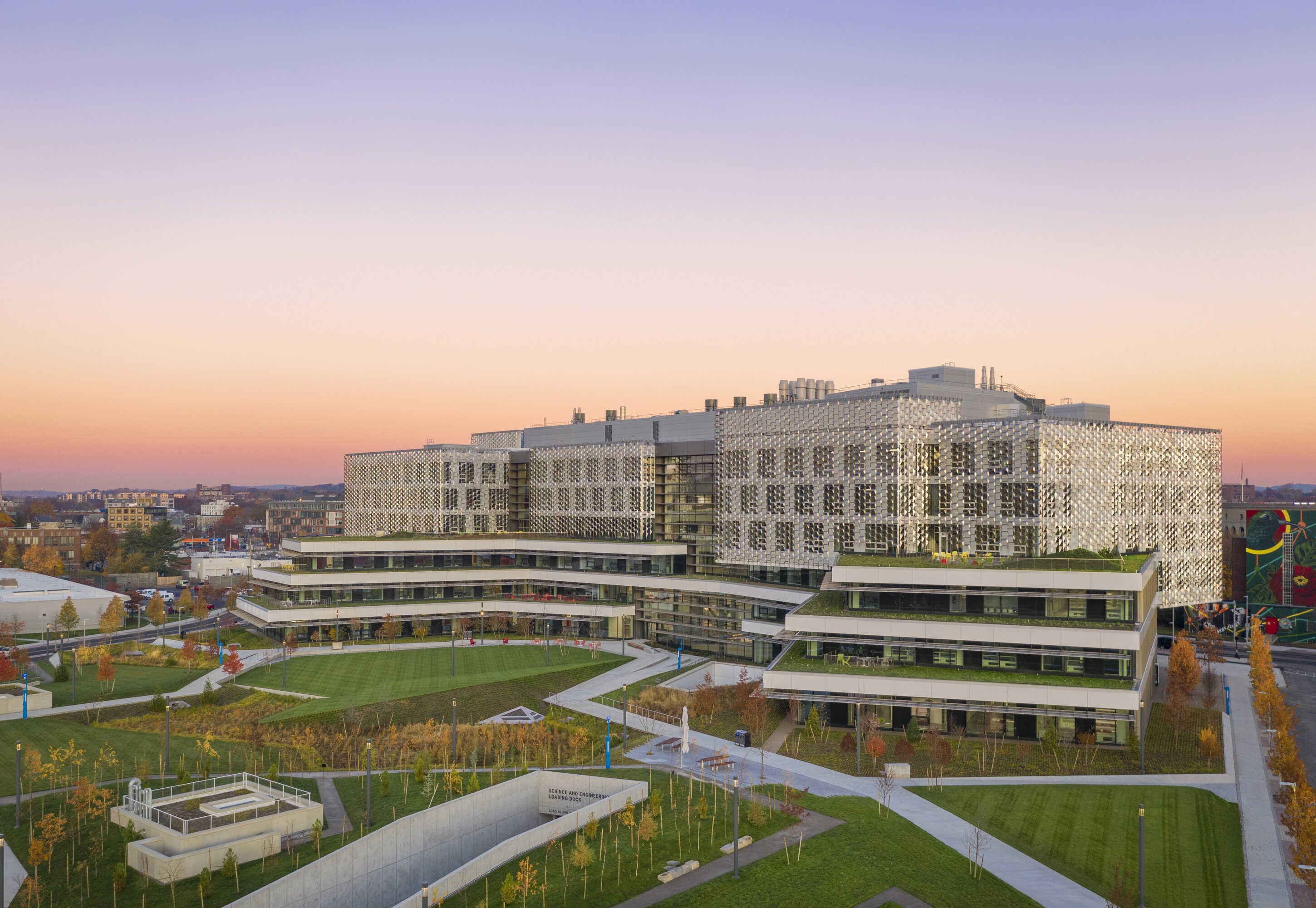 Project by Behnisch Architekturbuero, Harvard University Science and Engineering Complex (SEC). Large, modern campus building with terraced levels and patterned facade, surrounded by well-maintained lawns and paths in an evening atmosphere.