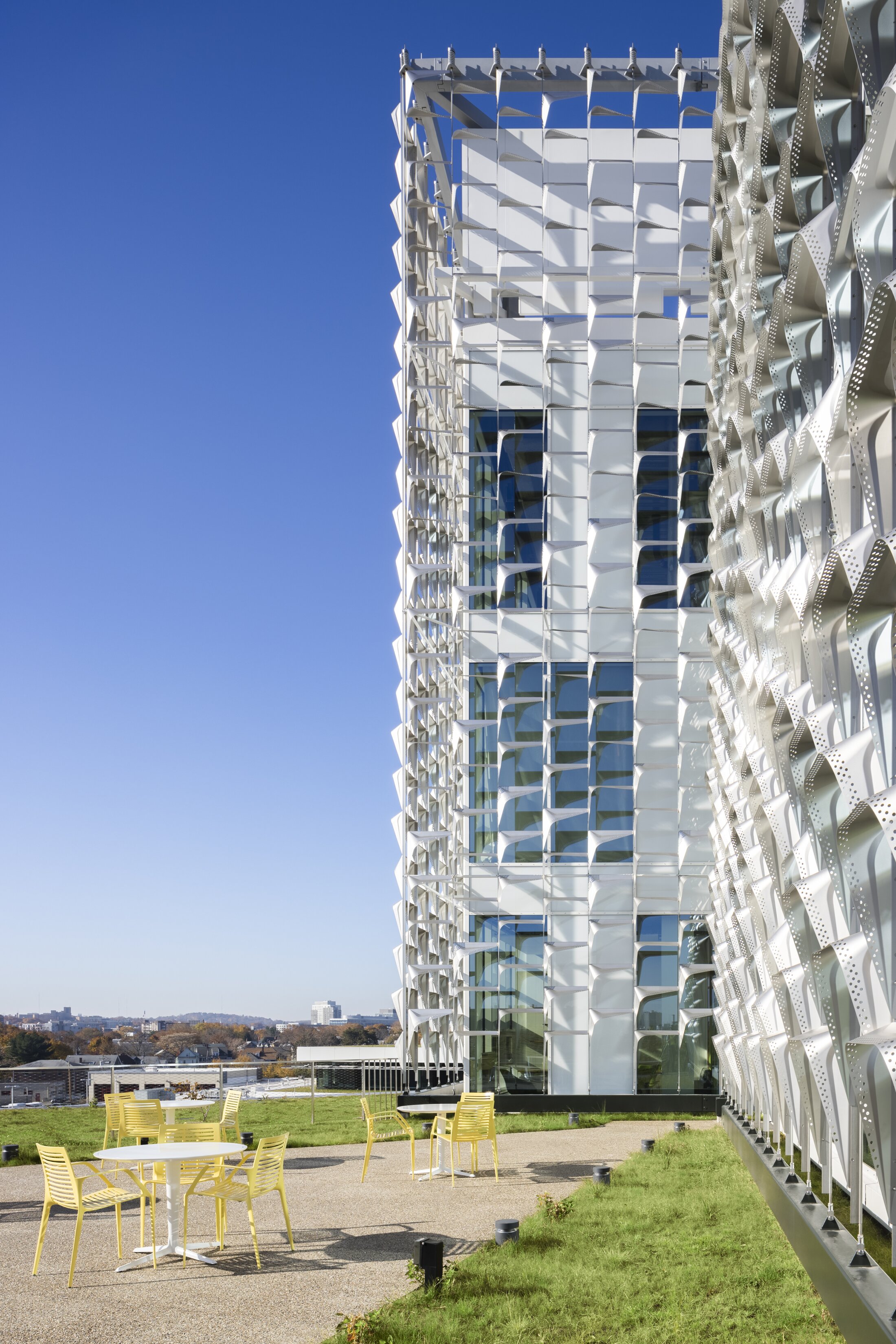 Project by Behnisch Architekturbuero, Harvard University Science and Engineering Complex (SEC). White facade with geometric sunshade panels on the modern building; yellow outdoor chairs and tables on a terrace with grass and city view.
