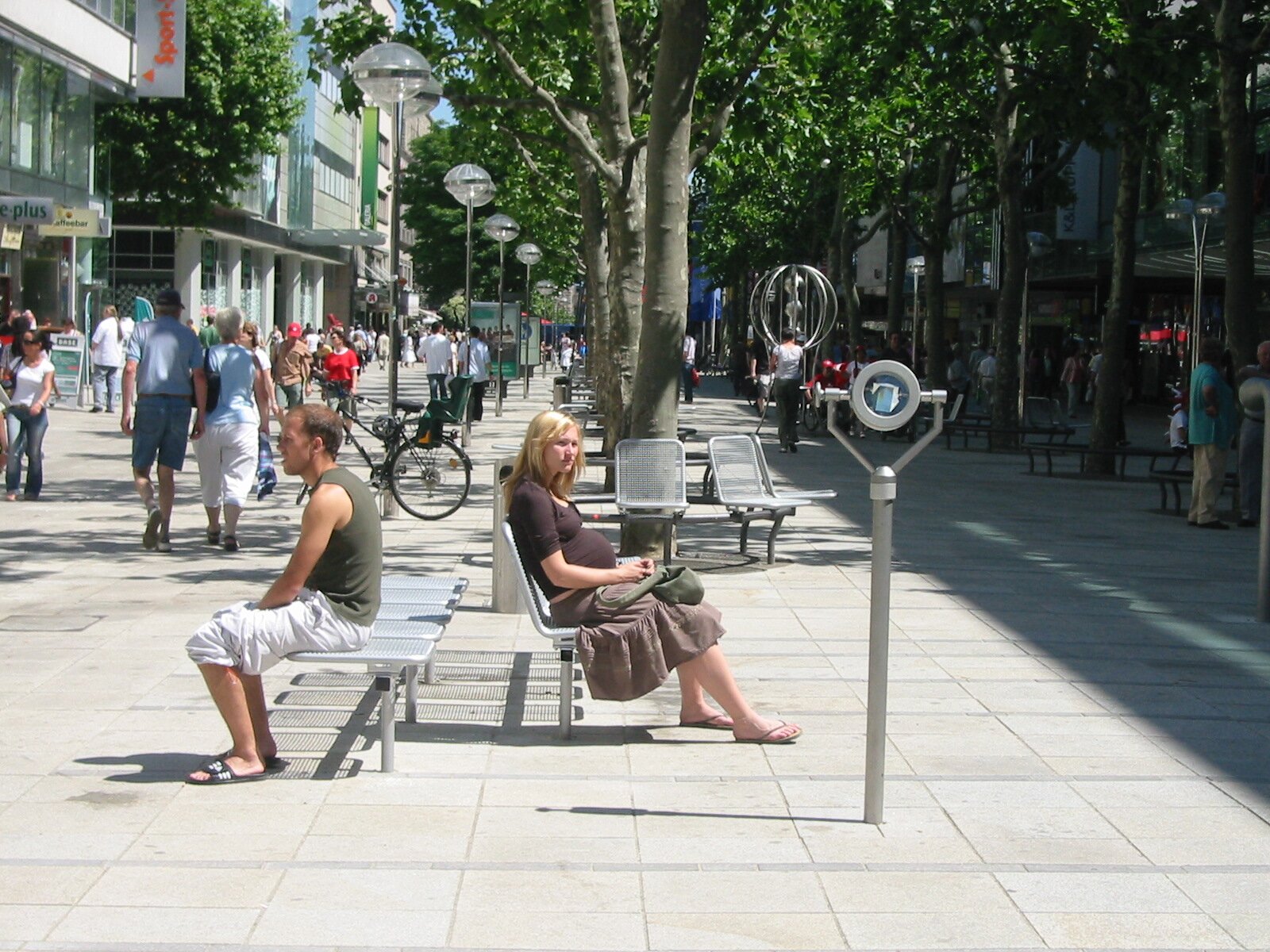 Project by Behnisch Architekturbuero, Refurbishment and Modernization K&ouml;nigsstra&szlig;e. People sit on benches along a tree-lined pedestrian street with shops, bicycles, and passersby in a busy urban setting.
