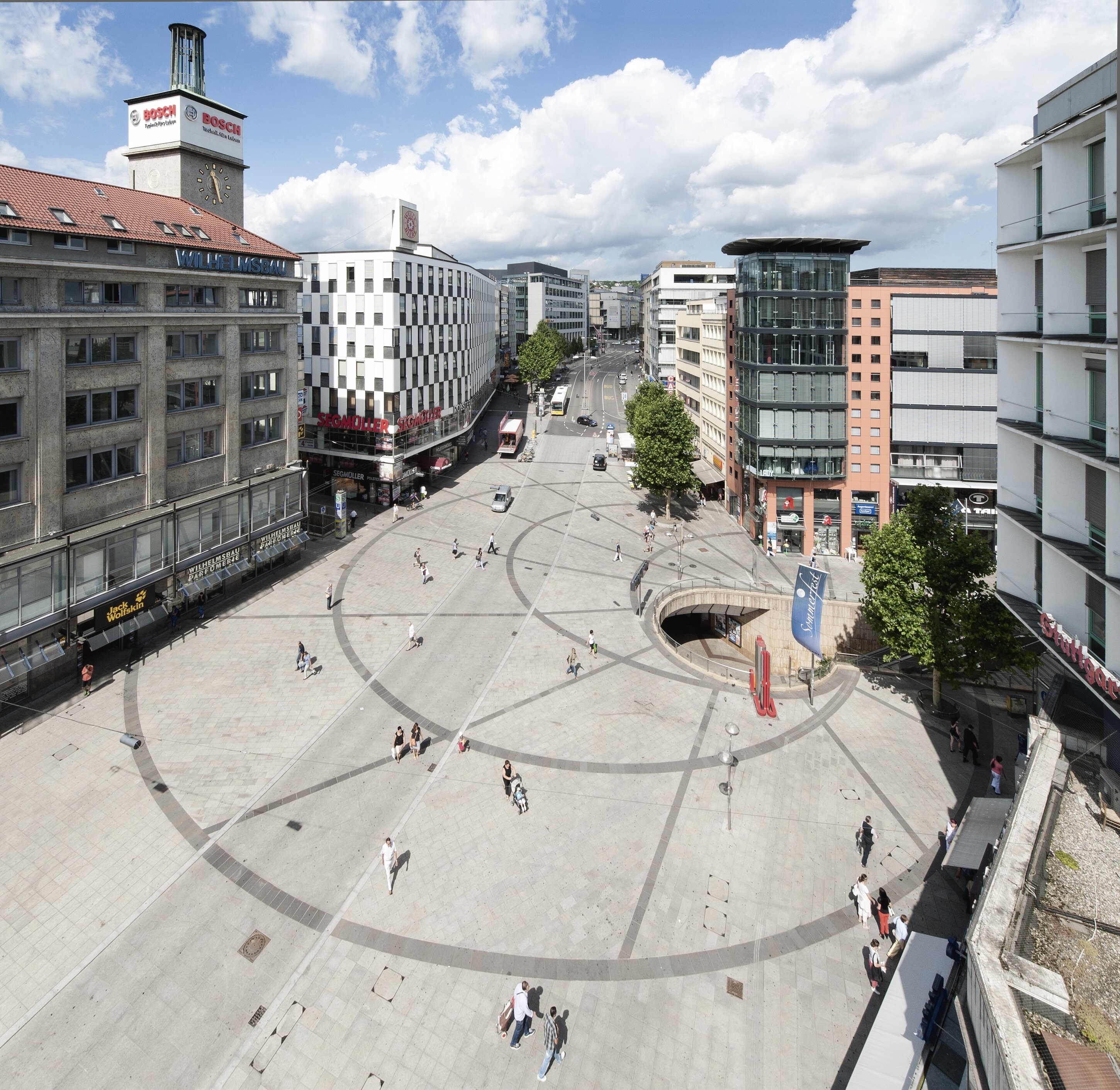 Project by Behnisch Architekturbuero, Refurbishment and Modernization K&ouml;nigsstra&szlig;e. Large city square in the city center of Stuttgart with a circular paving pattern, surrounded by medium-height buildings, pedestrians and a street that extends to the horizon.