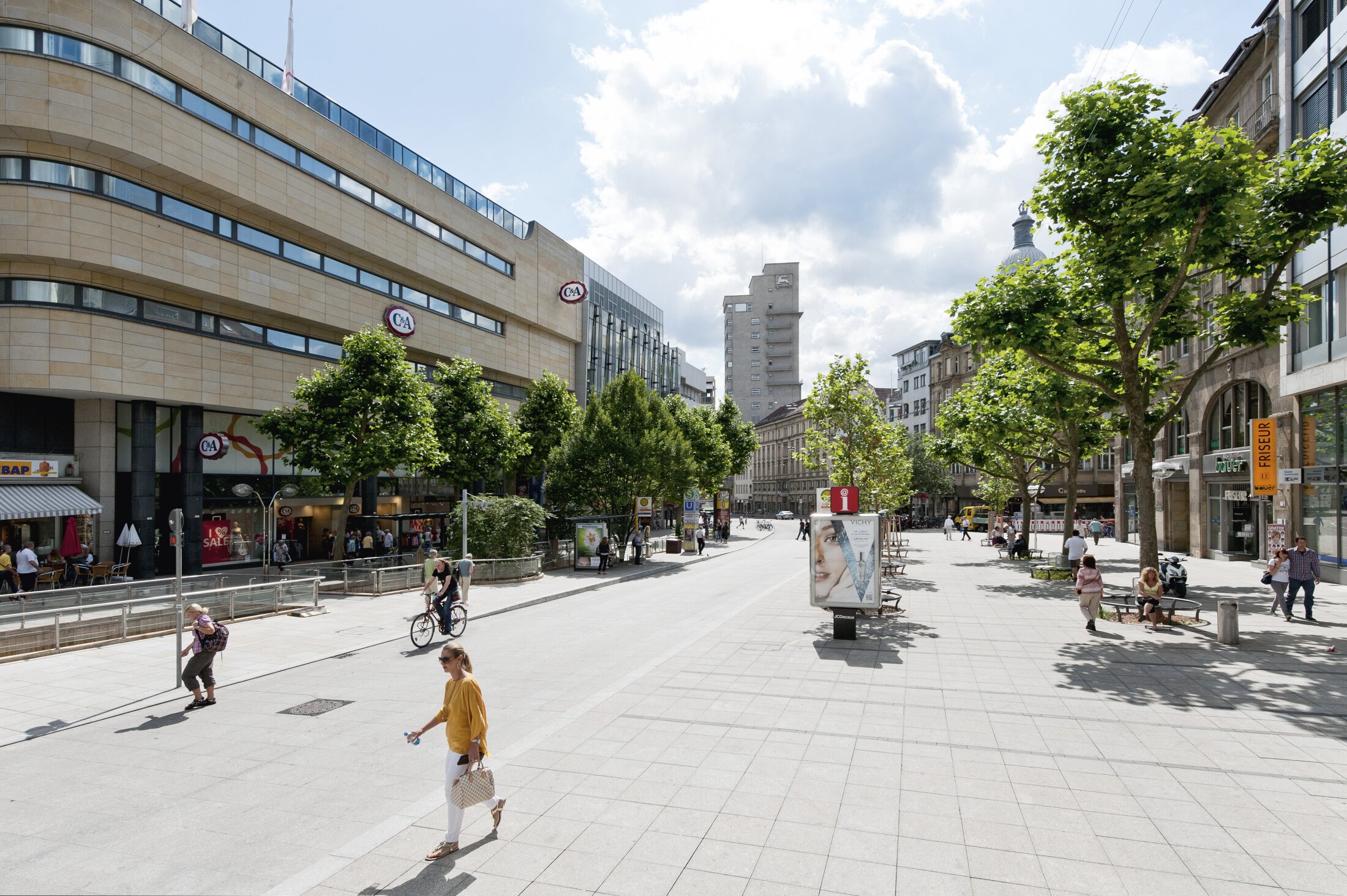 Project by Behnisch Architekturbuero, Refurbishment and Modernization K&ouml;nigsstra&szlig;e. Pedestrian street with trees, shops and cafes. People are walking and cycling on the road. Benches along a wide paved urban plaza under a cloudy sky.