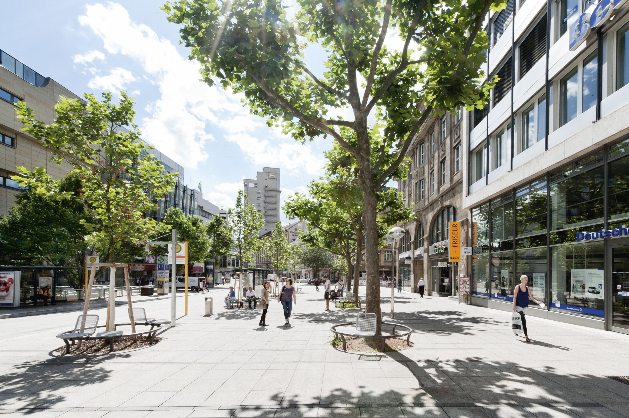 Project by Behnisch Architekturbuero, Refurbishment and Modernization K&ouml;nigsstra&szlig;e. Tree-lined pedestrian street with benches and shops. People are walking and sitting under a bright sky with scattered clouds.