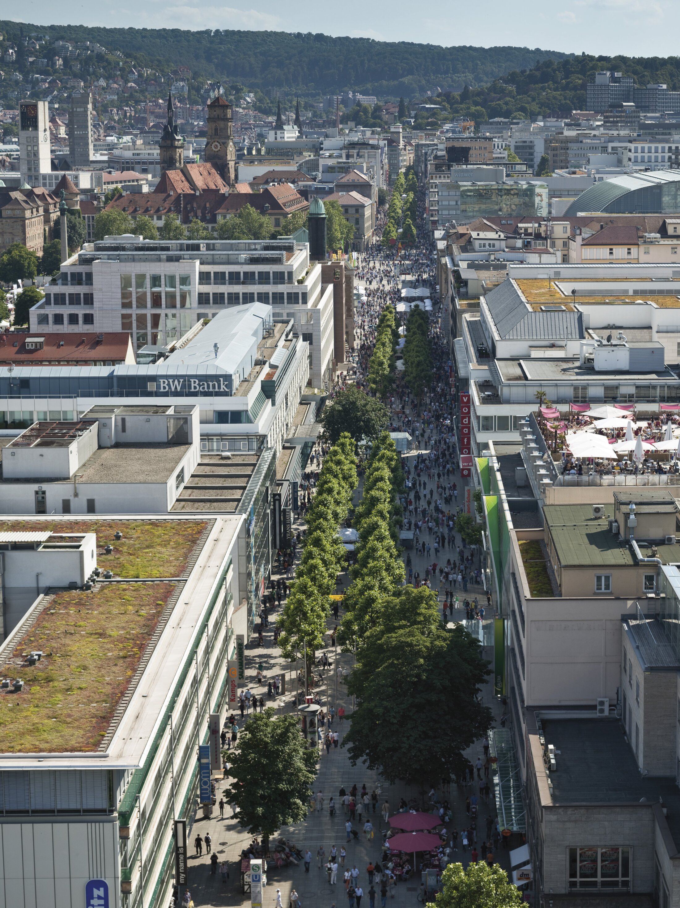 Project by Behnisch Architekturbuero, Refurbishment and Modernization K&ouml;nigsstra&szlig;e. Aerial view of the busy pedestrian street lined with trees and buildings, filled with crowds, with hills and cityscape in the background.