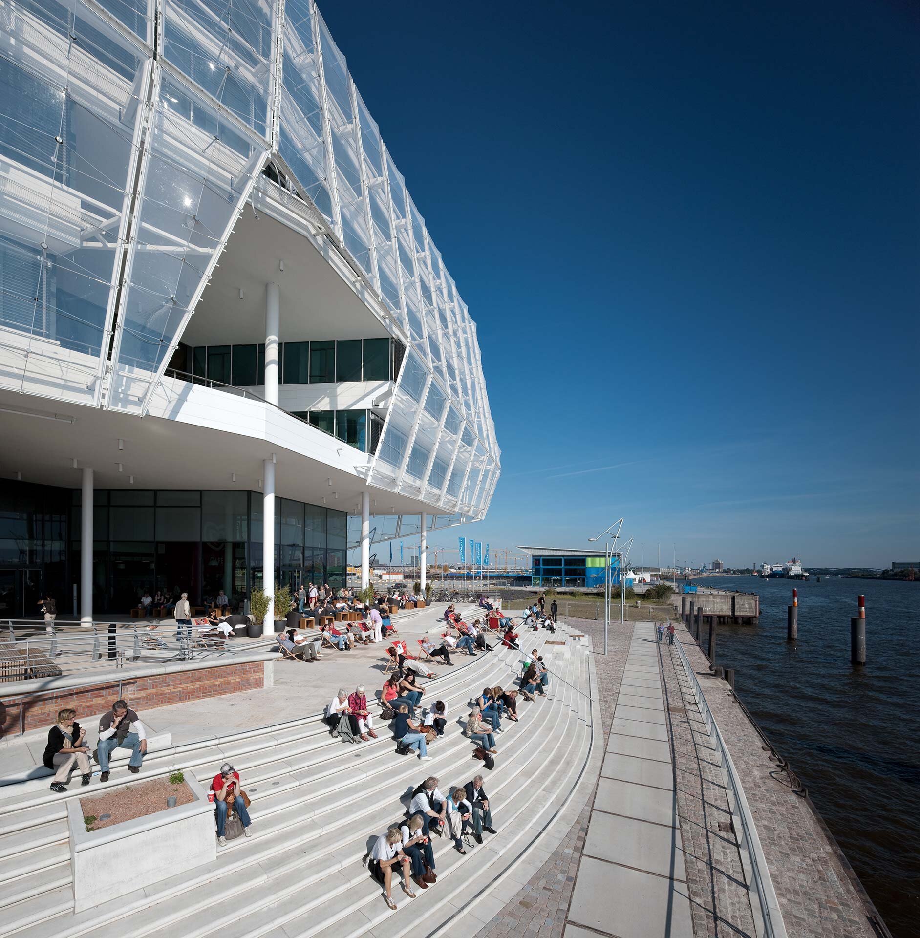 Project by Behnisch Architekturbuero, Unilever Headquarters for Germany, Austria, and Switzerland. People sitting on stepped terraces beside the waterfront, next to the modern building, with a promenade and river under a clear blue sky.