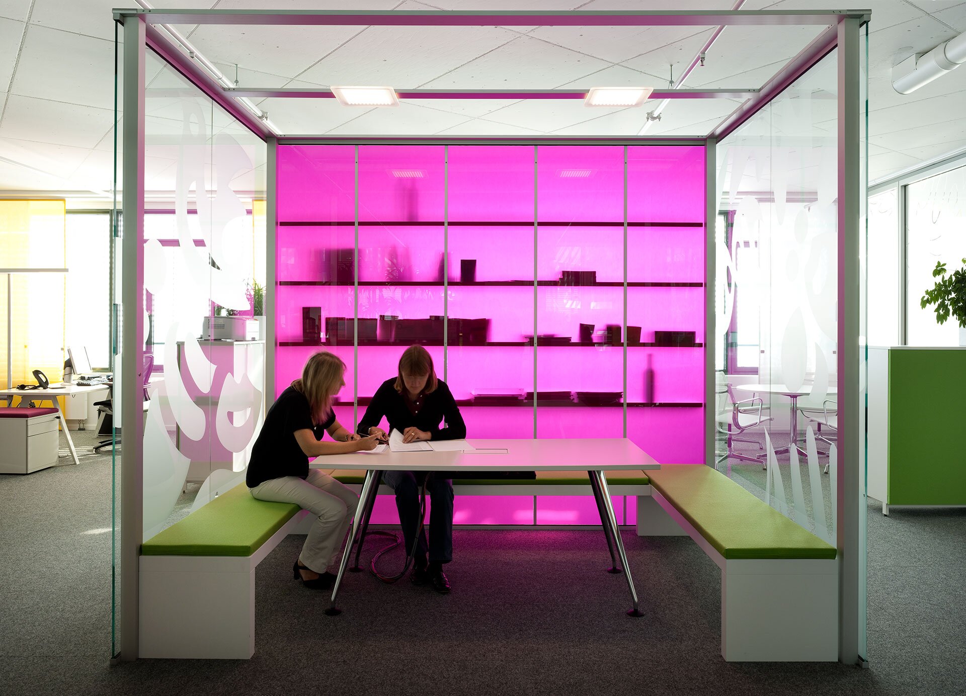 Project by Behnisch Architekturbuero, Unilever Headquarters for Germany, Austria, and Switzerland. Two people seated at a table in a glass meeting booth with green benches and a bright pink backlit wall of shelves behind them.
