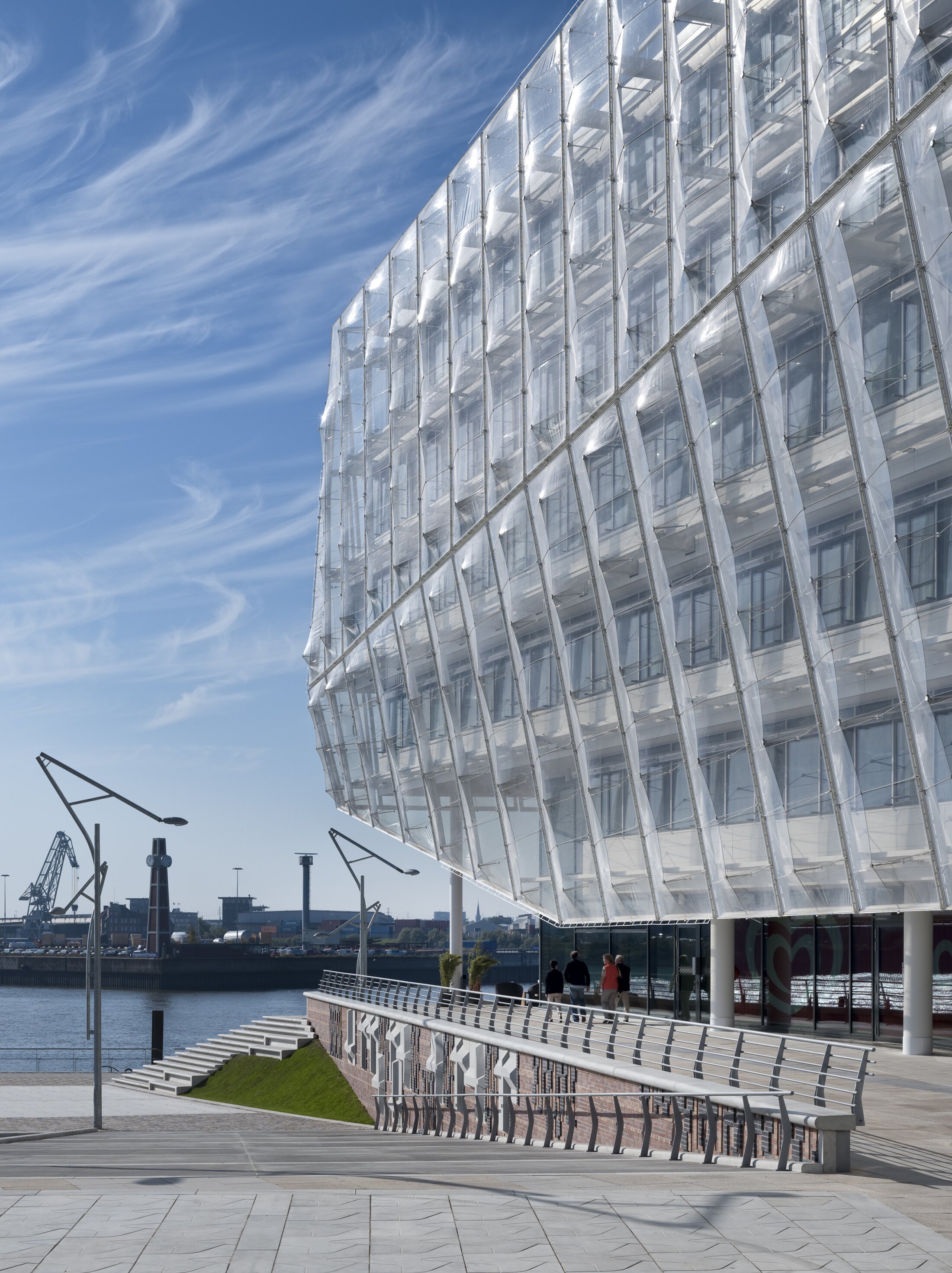 Project by Behnisch Architekturbuero, Unilever Headquarters for Germany, Austria, and Switzerland. Close-up of the ETFE membrane facade of the waterfront building, with a promenade, stairs and some strollers under a blue sky.