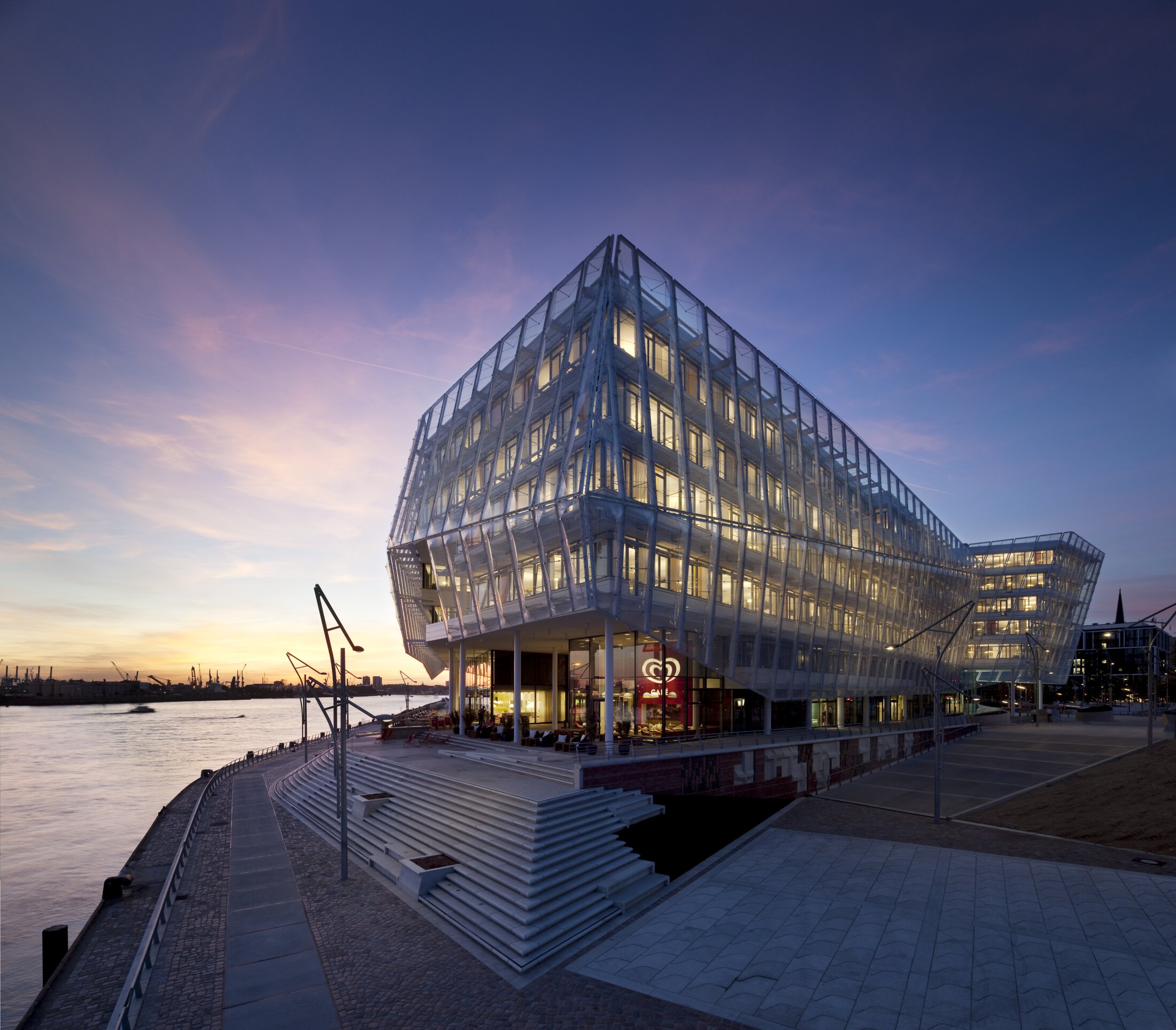 Project by Behnisch Architekturbuero, Unilever Headquarters for Germany, Austria, and Switzerland. Illuminated glass building by the river at dusk, with stepped terraces, waterfront promenade, and pastel sunset sky.