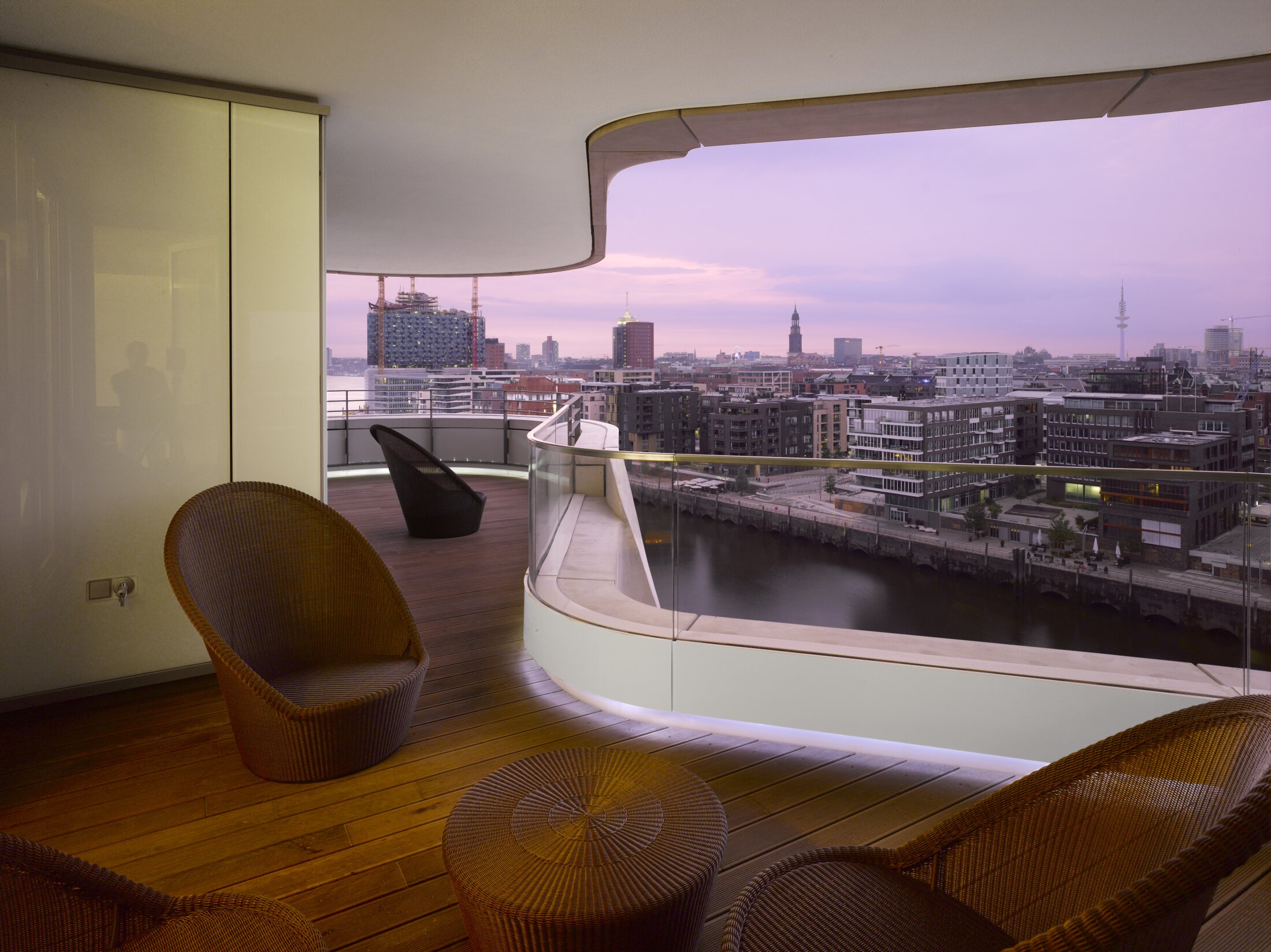 Project by Behnisch Architekturbuero, Marco Polo Tower. Curved balcony with wicker chairs overlooking the river and modern city skyline at dusk, viewed from a furnished interior terrace.
