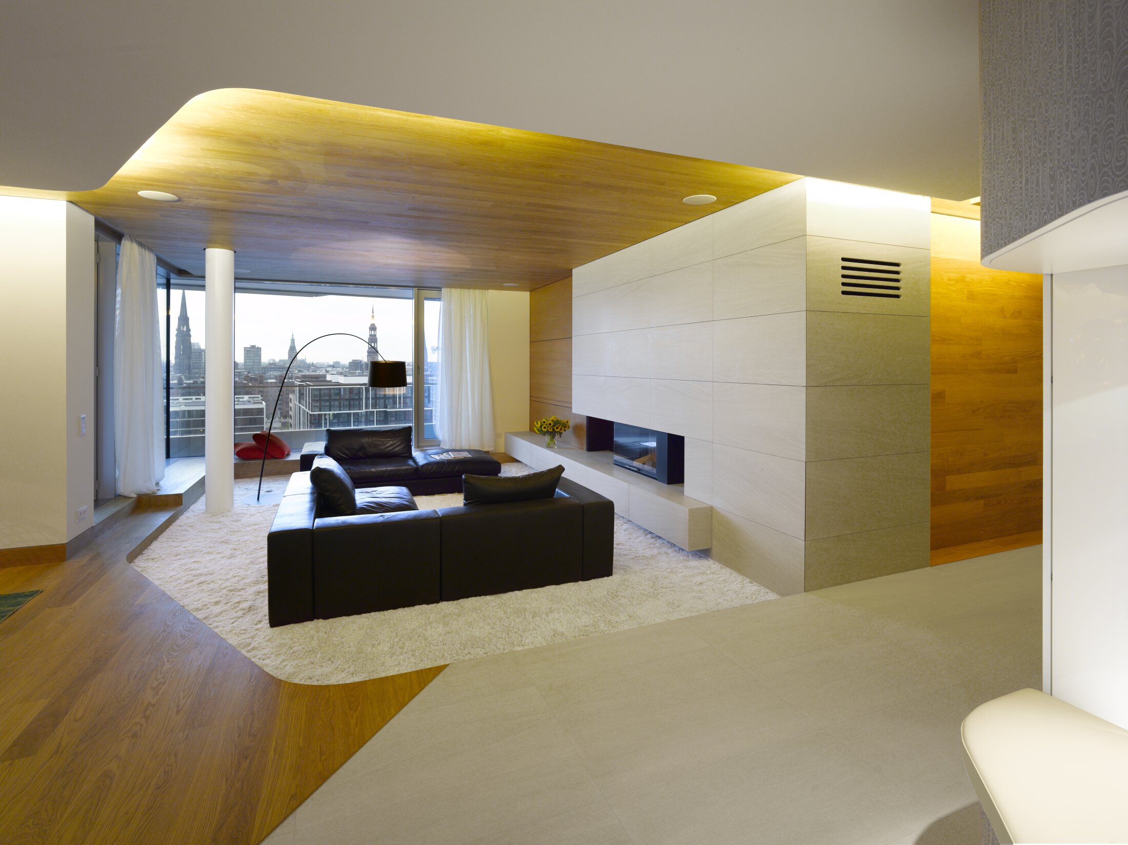 Project by Behnisch Architekturbuero, Marco Polo Tower. Modern living room with black leather sofas, fireplace, wood ceiling, and large window overlooking a city skyline.