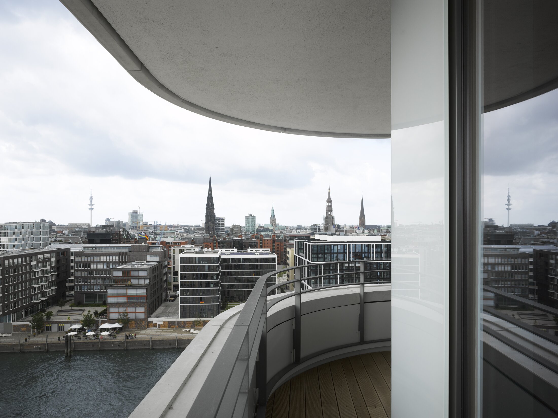 Project by Behnisch Architekturbuero, Marco Polo Tower. Curved balcony with glass railing overlooking the river and city skyline with church spires and towers under cloudy sky.